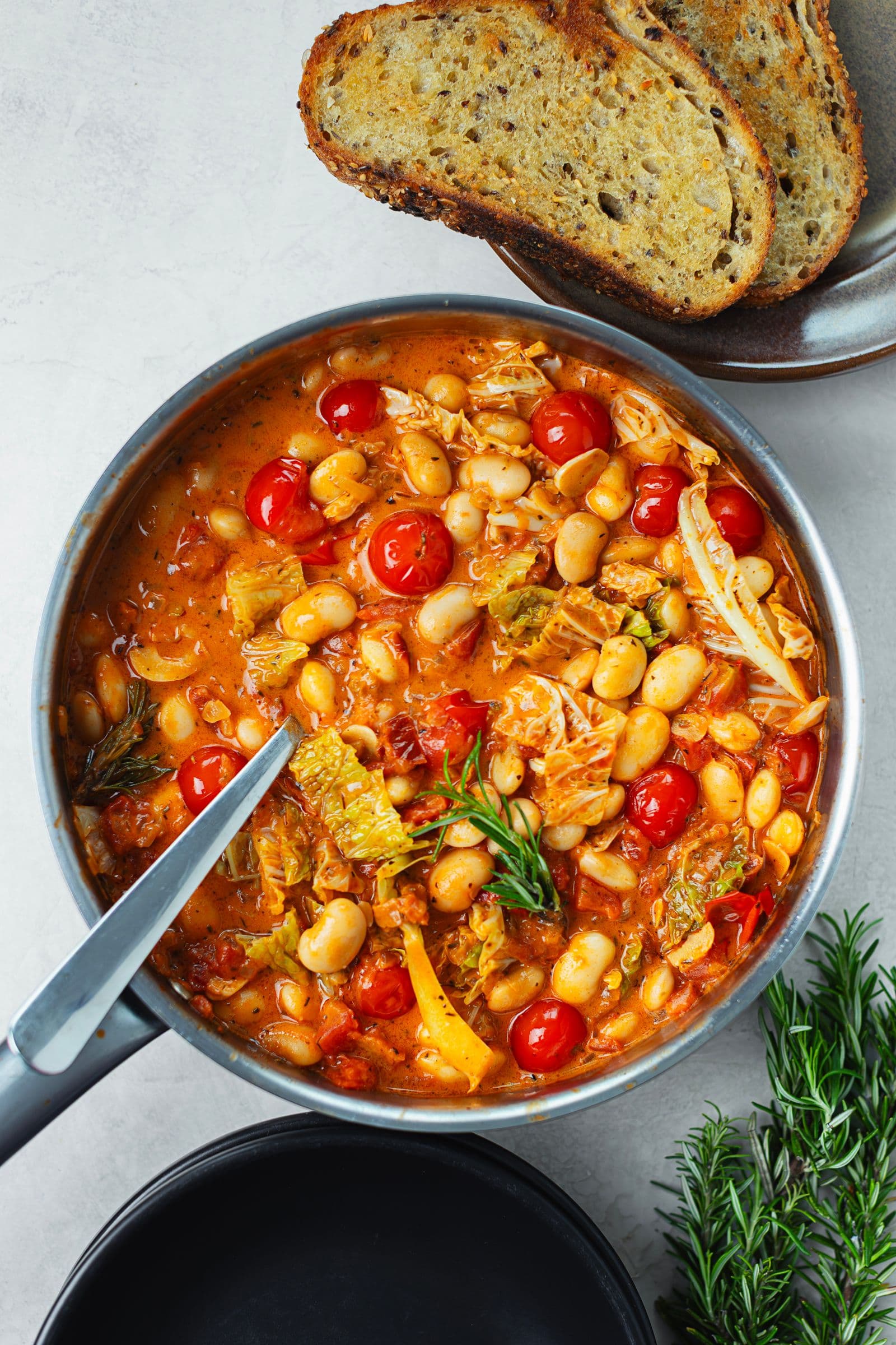Overhead view of the stew in a pot with visible beans, tomatoes and cabbage, served next to slices of toasted bread.