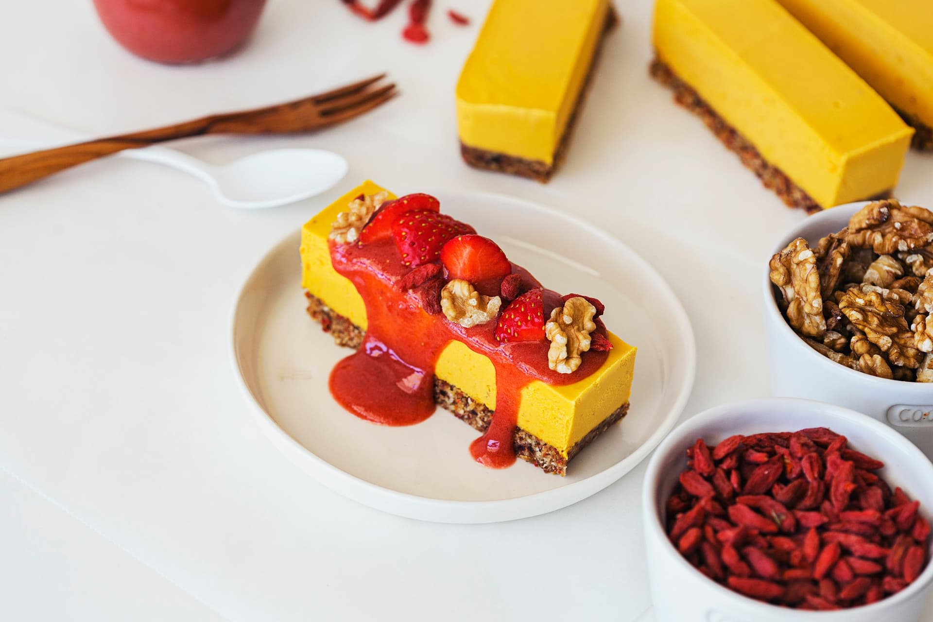 Slice of orange, goji, and strawberry raw vegan cheesecake topped with fresh berries and sauce.