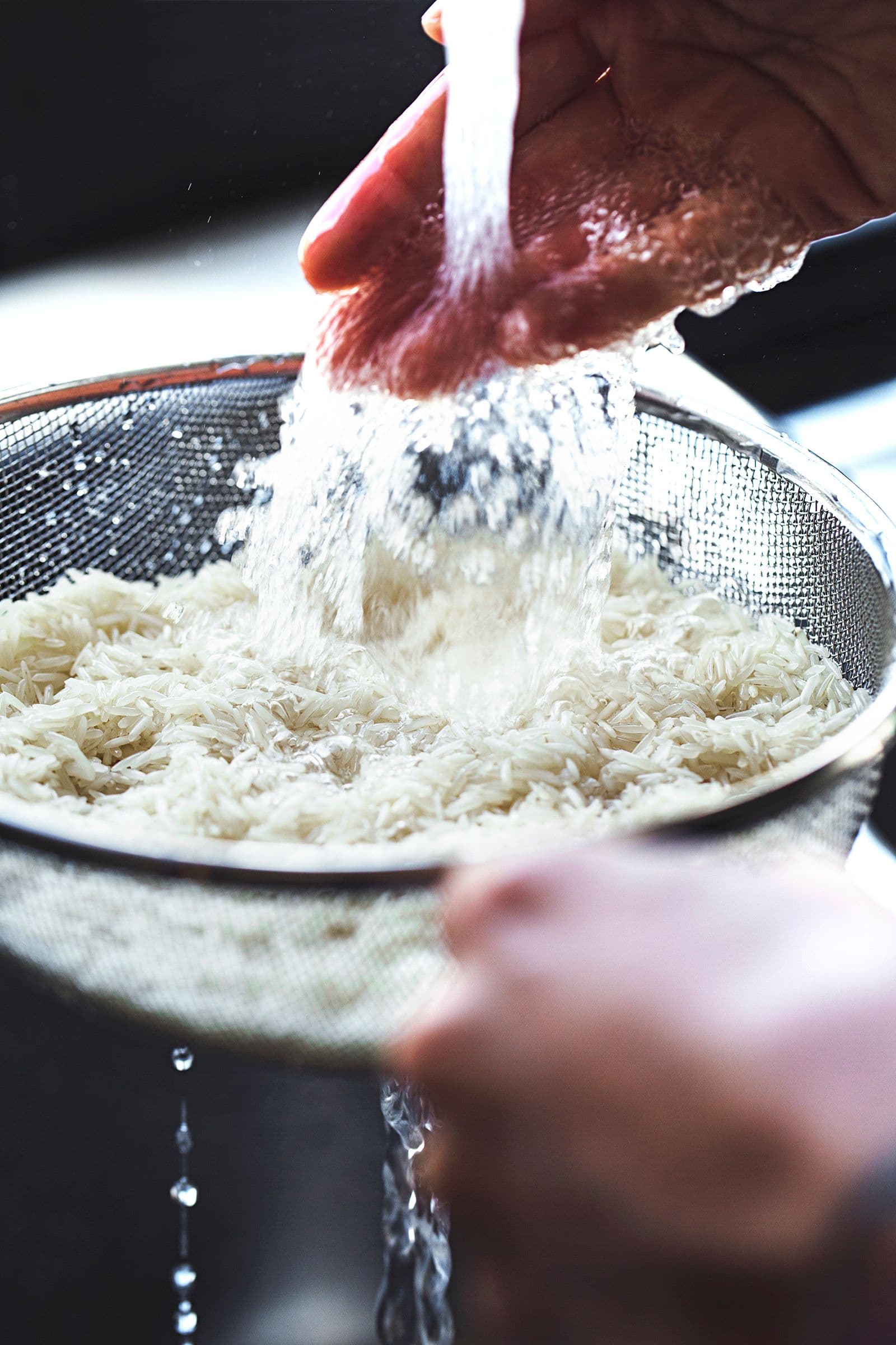 Mãos a lavar arroz basmati em água corrente num coador de malha fina.