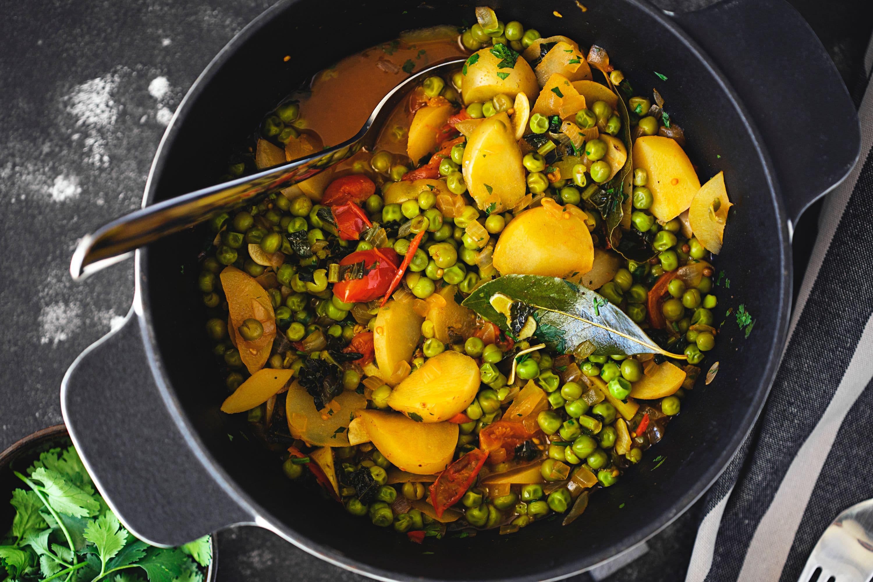 Vista aproximada de ensopado vegan de ervilhas num tacho preto com batatas, tomates-cereja e louro.