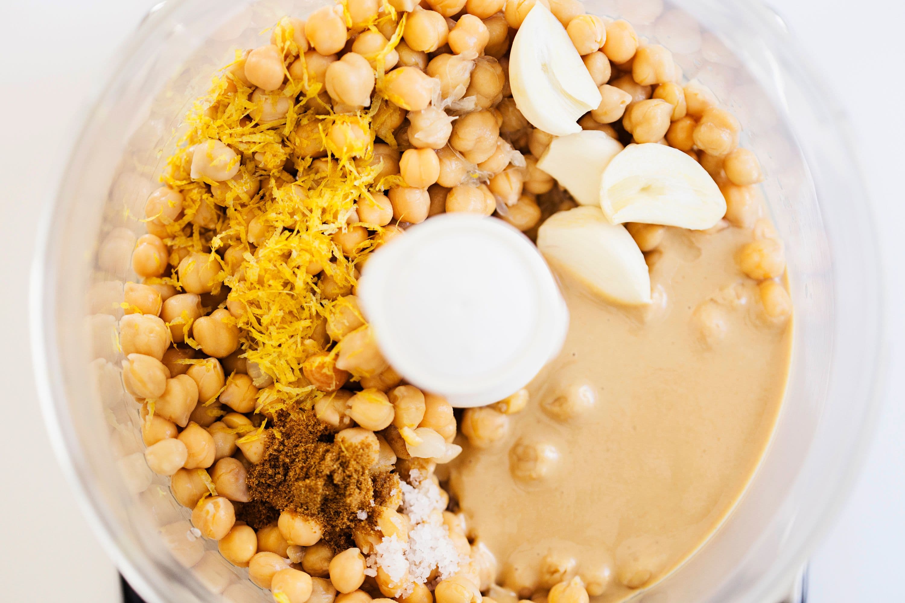 Food processor bowl filled with chickpeas, tahini, lemon zest, garlic cloves, and spices.