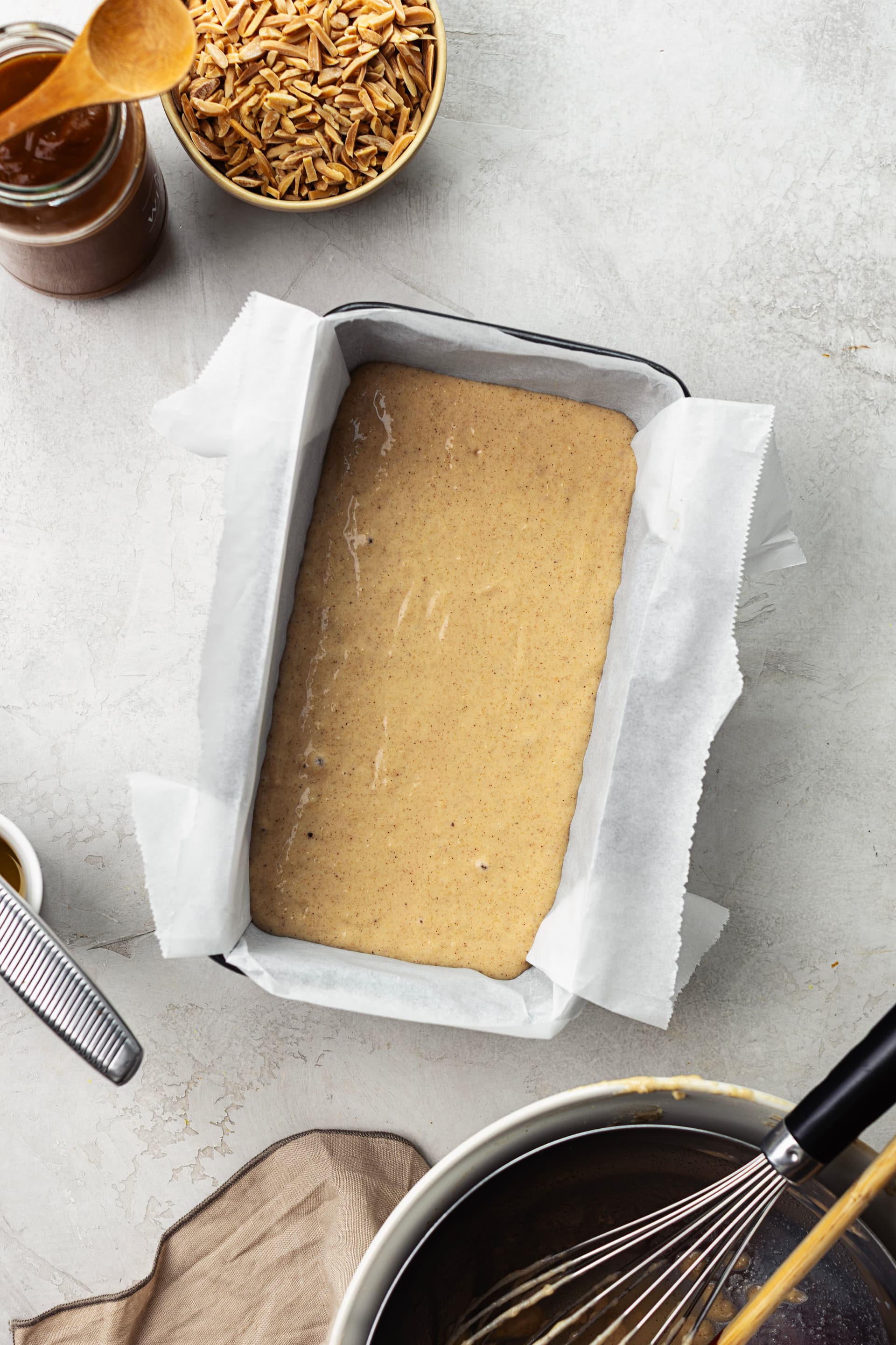 Massa de bolo de amêndoa crua numa forma rectangular forrada com papel vegetal, com caramelo de coco e amêndoas palitadas ao fundo.