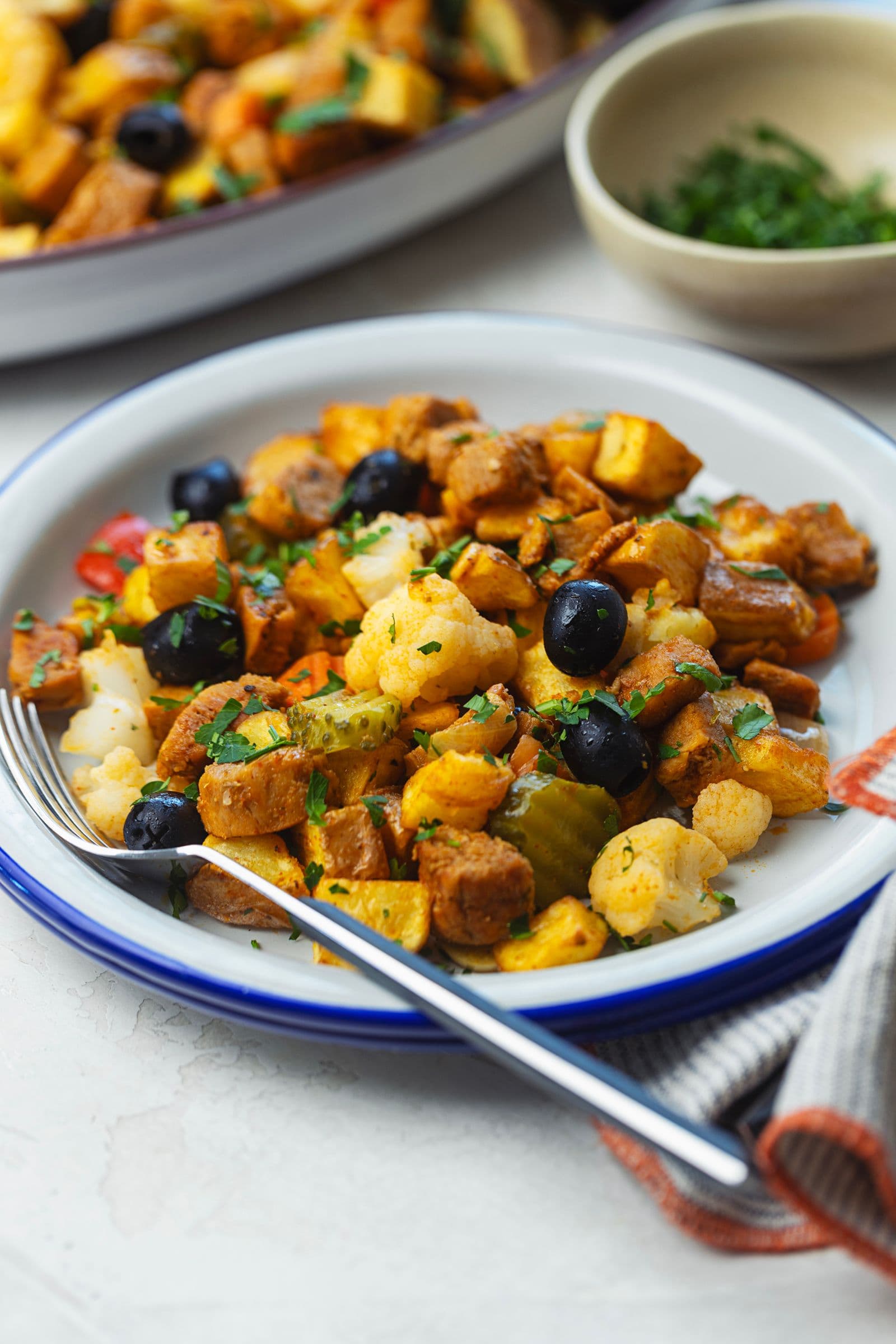 Prato de borda azul com porção de pica pau de soja em ângulo frontal ligeiramente elevado, com azeitonas pretas, pickles de couve-flor, batatas e soja caramelizada polvilhados com salsa