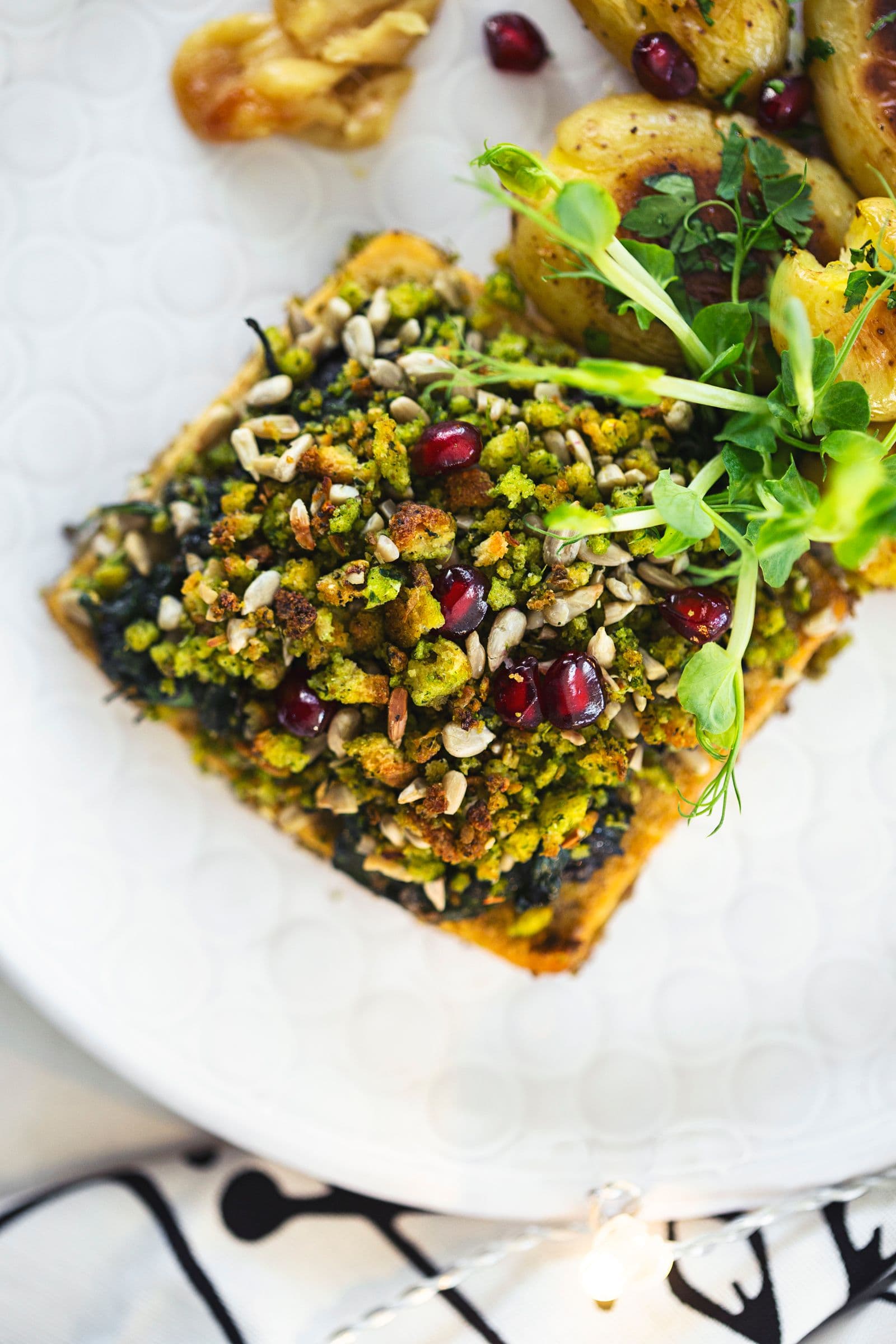 A piece of tofu roast topped with crumble served with roasted potatoes and pea shoots.