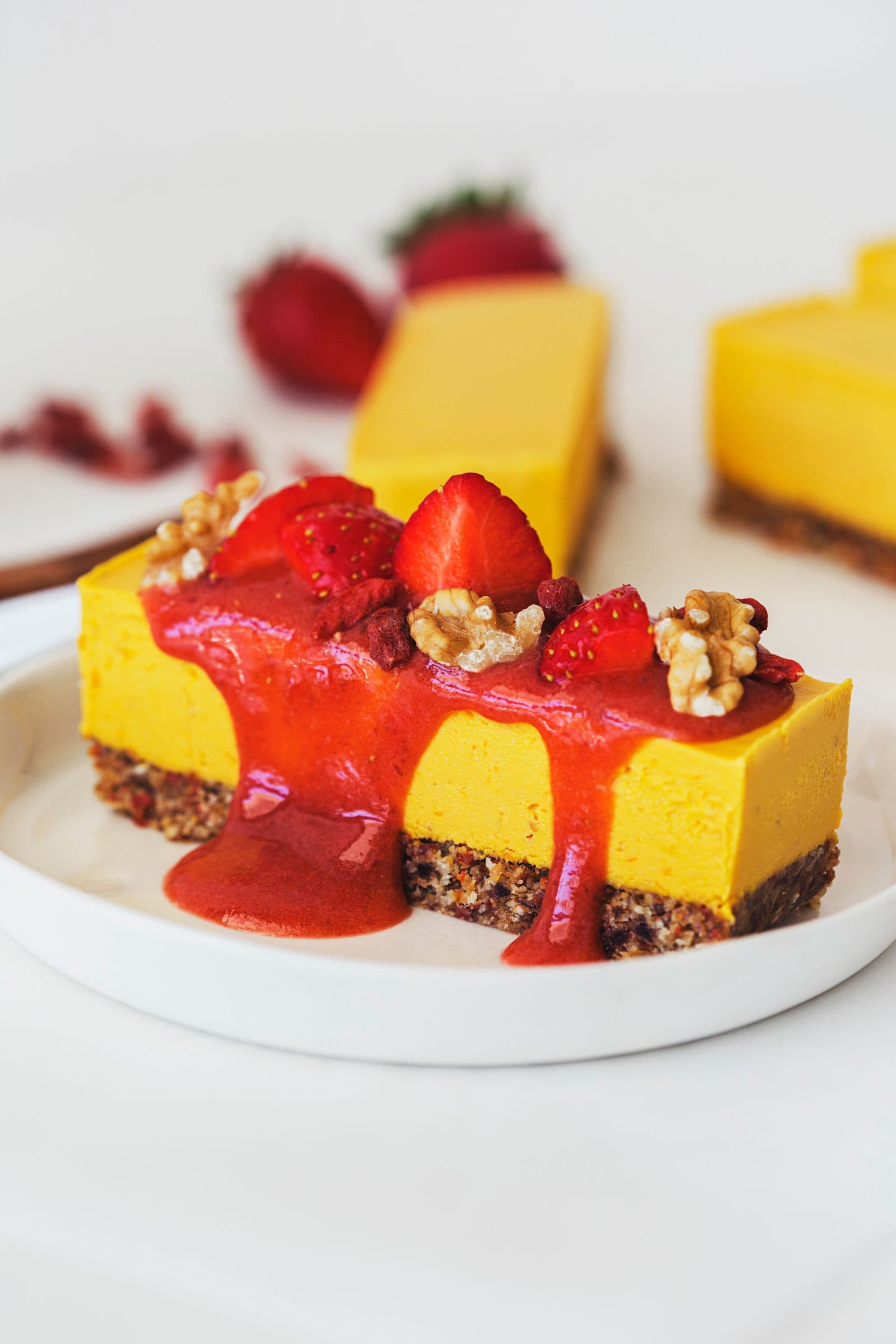 Slice of orange, goji, and strawberry raw vegan cheesecake topped with fresh strawberries, walnuts and sauce.
