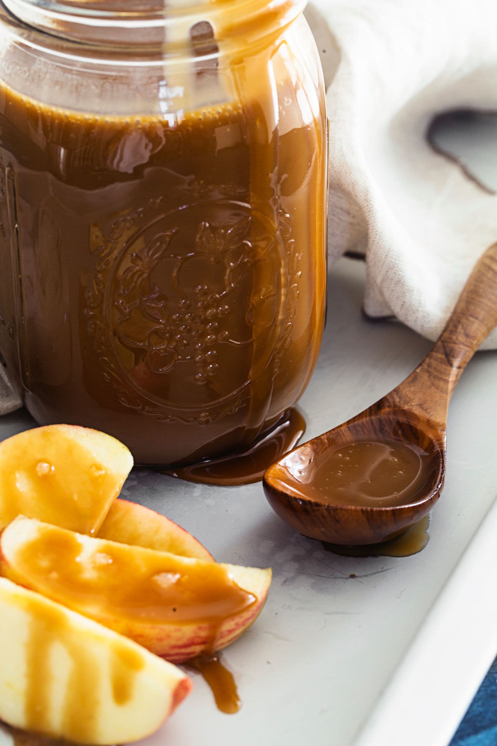Molho de caramelo derramado sobre fatias de maçã, com frasco e colher de madeira em fundo claro.