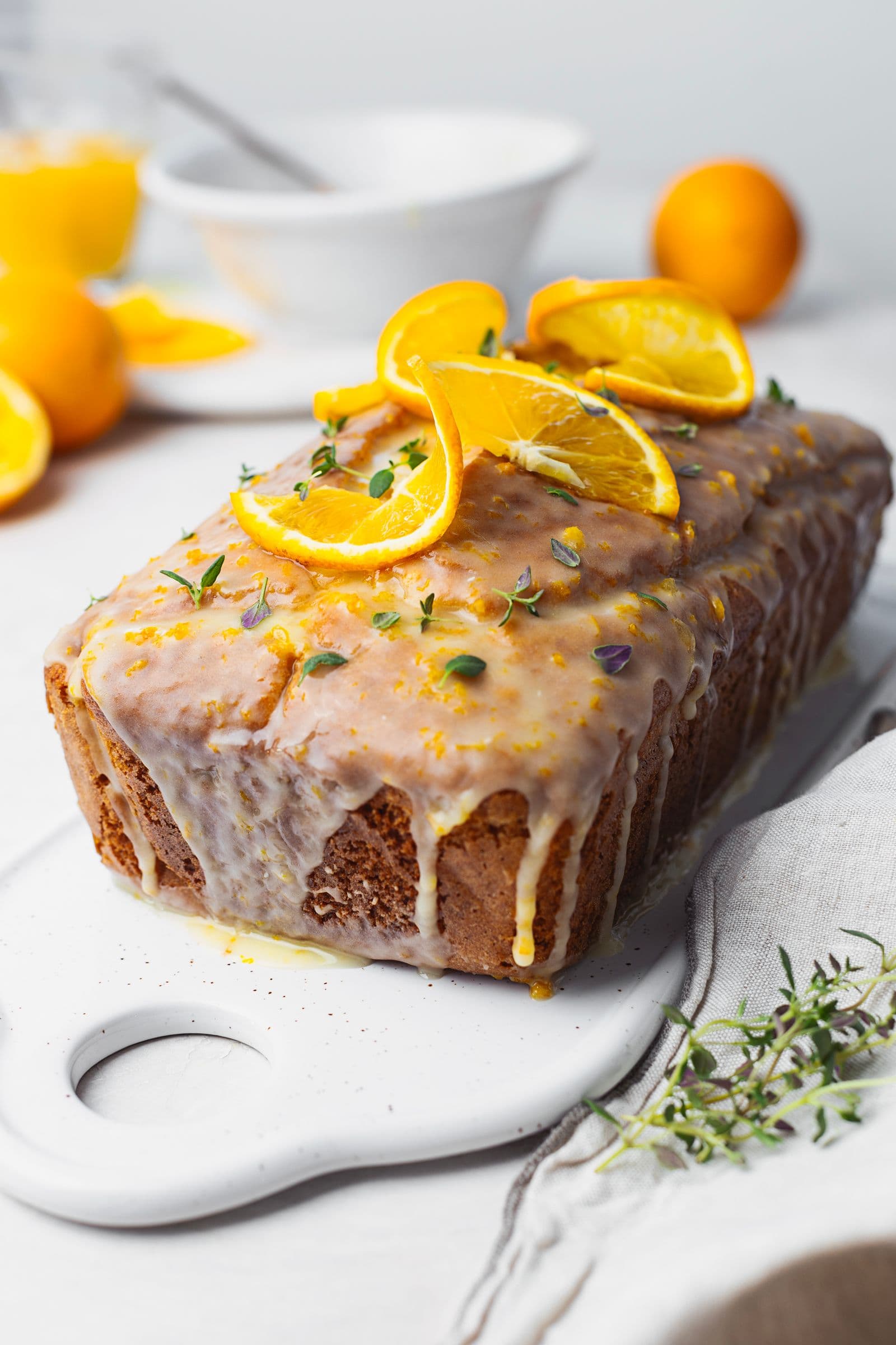 Bolo de laranja com calda a escorrer pelas laterais, decorado com rodelas de laranja e tomilho fresco, sobre uma travessa branca.