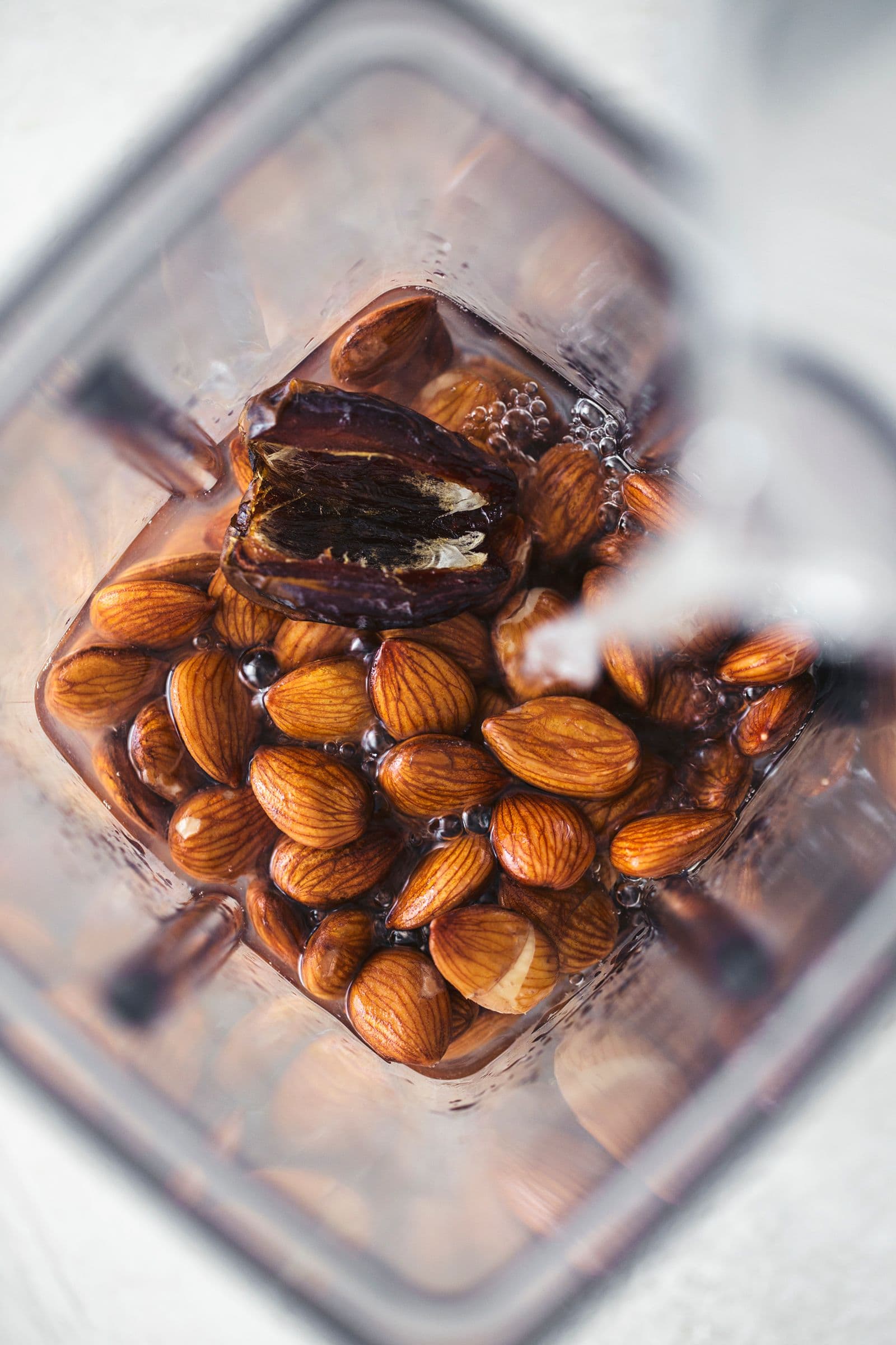Top view of a blender jar with raw almonds, a date and water being poured in.