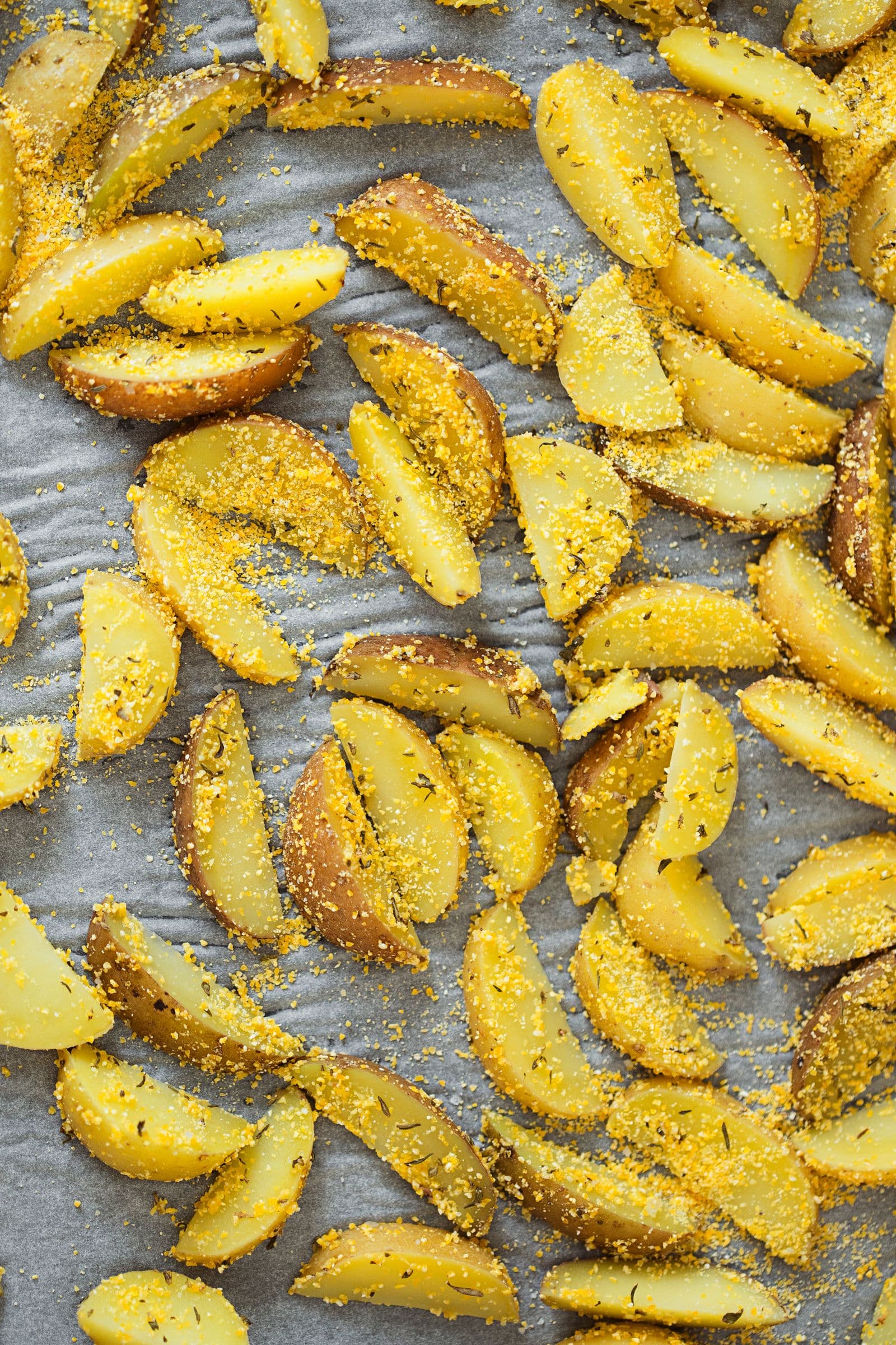 Raw potato wedges coated in herbs and polenta, arranged on a lined baking tray.