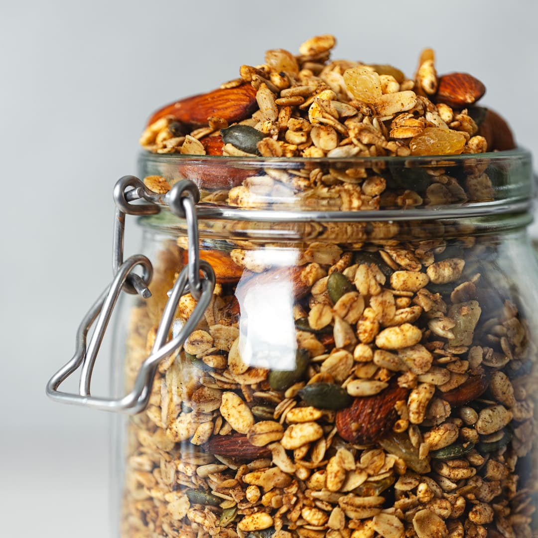 Granola crocante com especiarias chai, amêndoas e sementes num frasco de vidro, com um pouco espalhada na mesa.