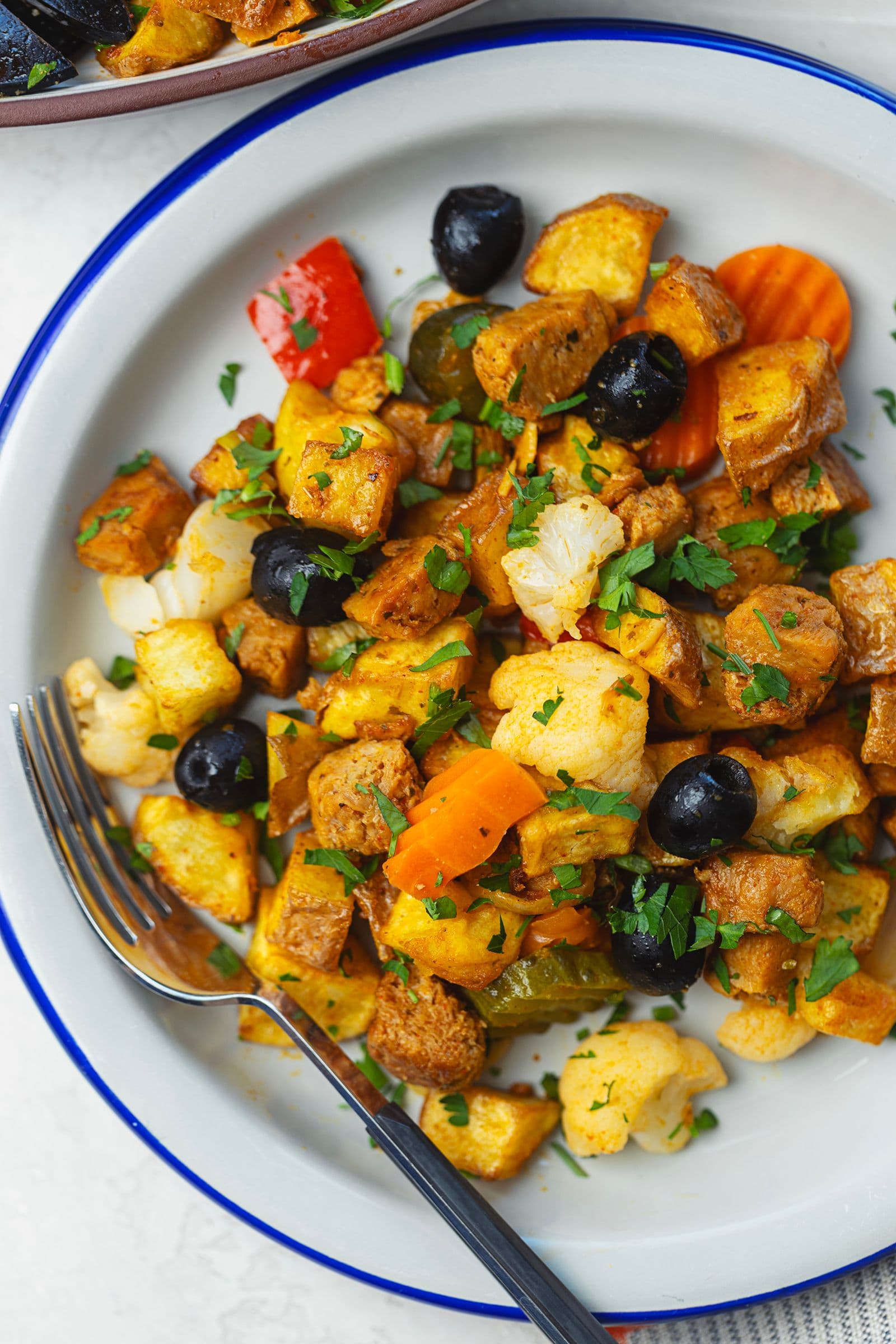 Overhead view of blue-rimmed plate with a serving of soy pica pau: fried potatoes, soy chunks, black olives, cauliflower, pepper, carrot and fresh parsley, with a fork