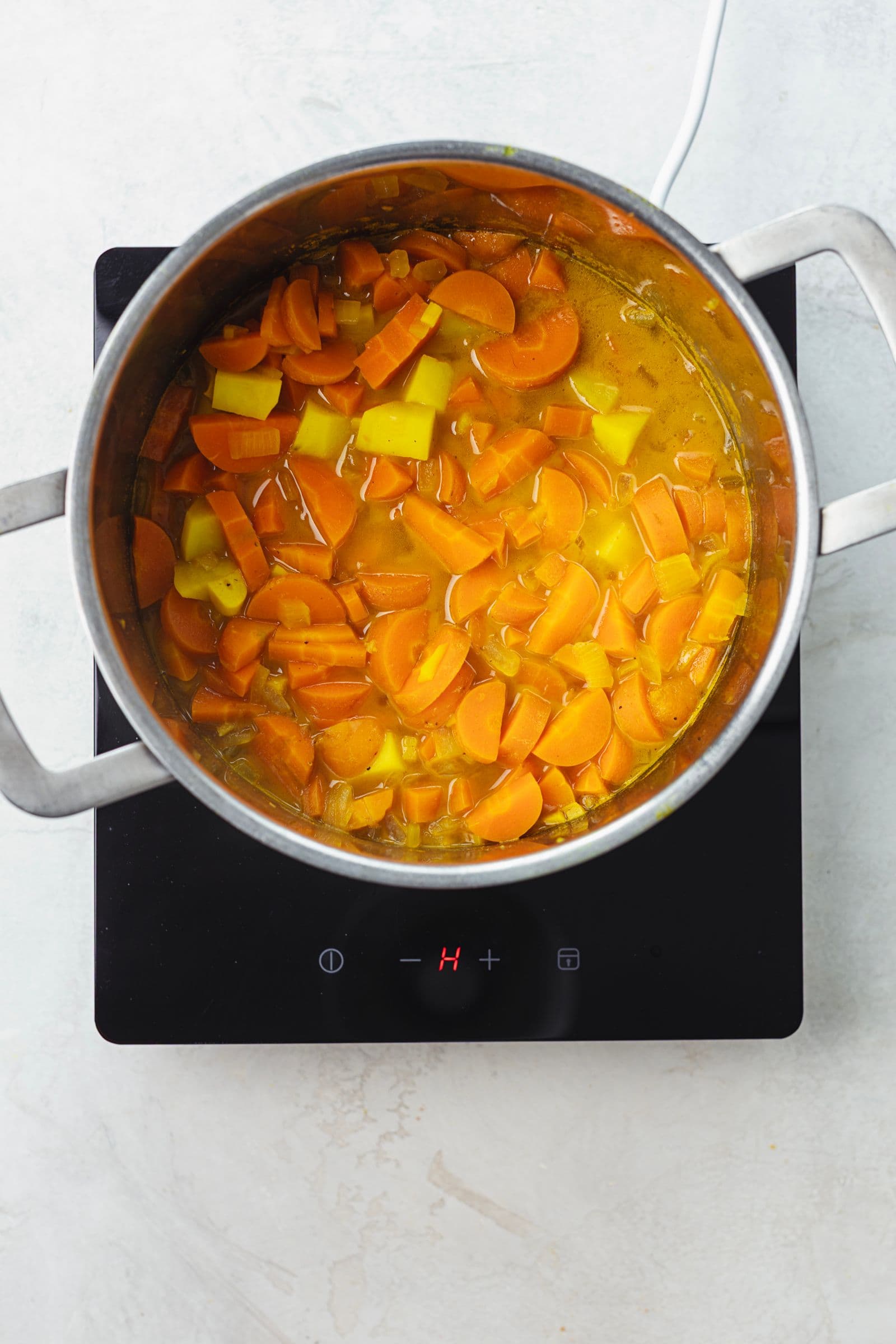 Pot on the stove with carrots, potatoes, and onions simmering in golden broth, overhead view of the soup cooking process.