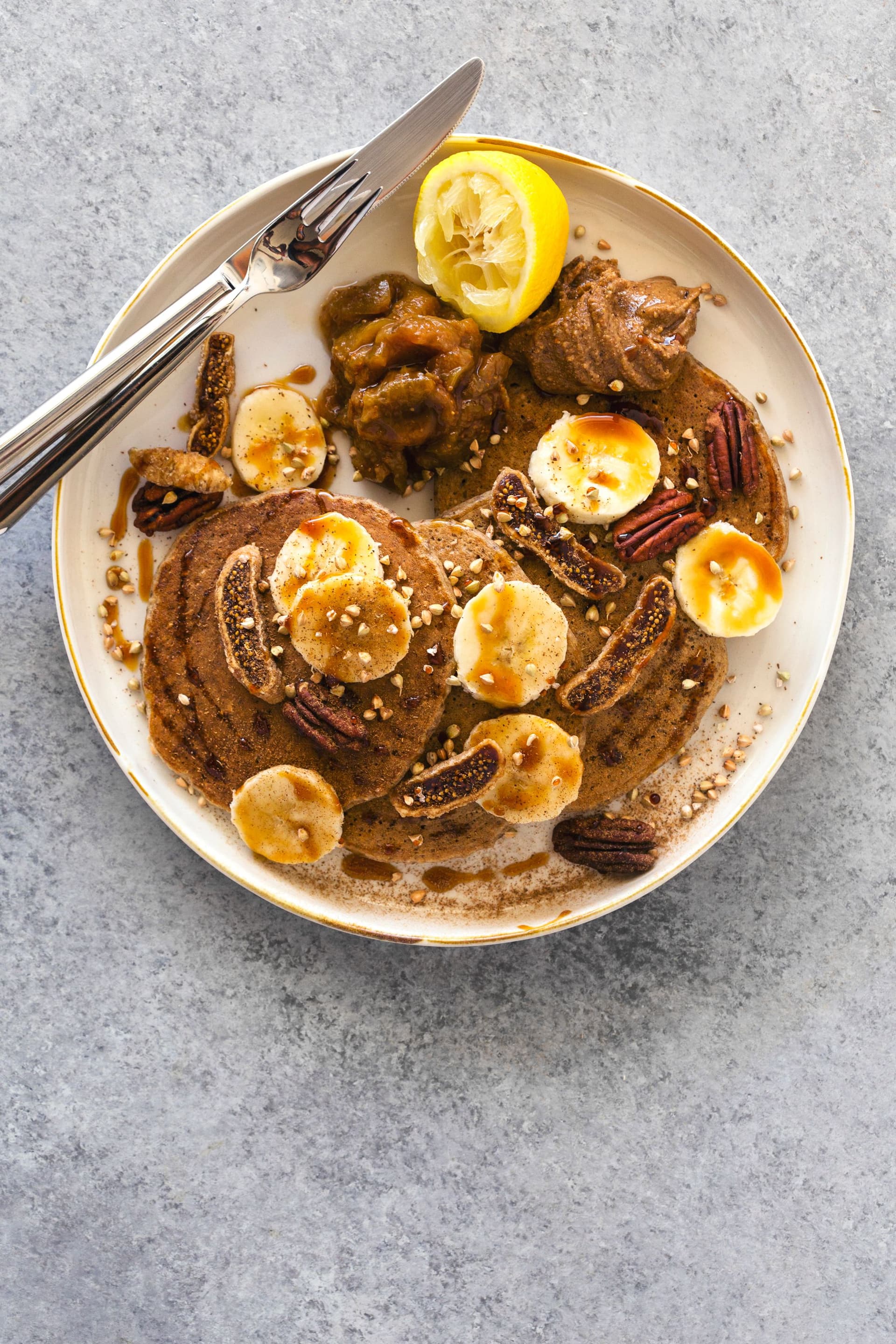 Panquecas de banana e limão servidas com compota, banana e nozes pecã, prontas a comer com talheres.