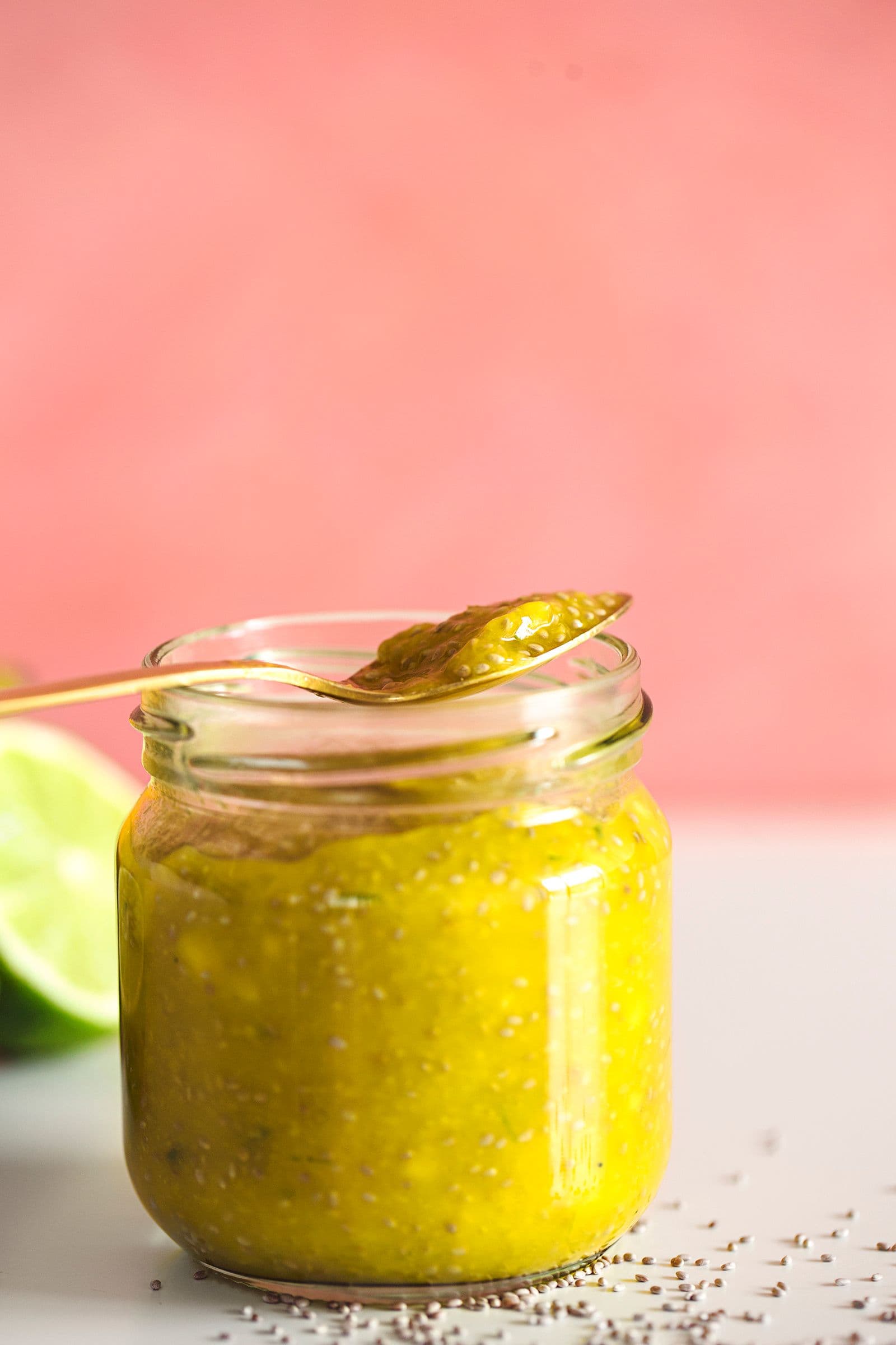 Spoon resting atop a jar of quick mango chia jam on a pink background.