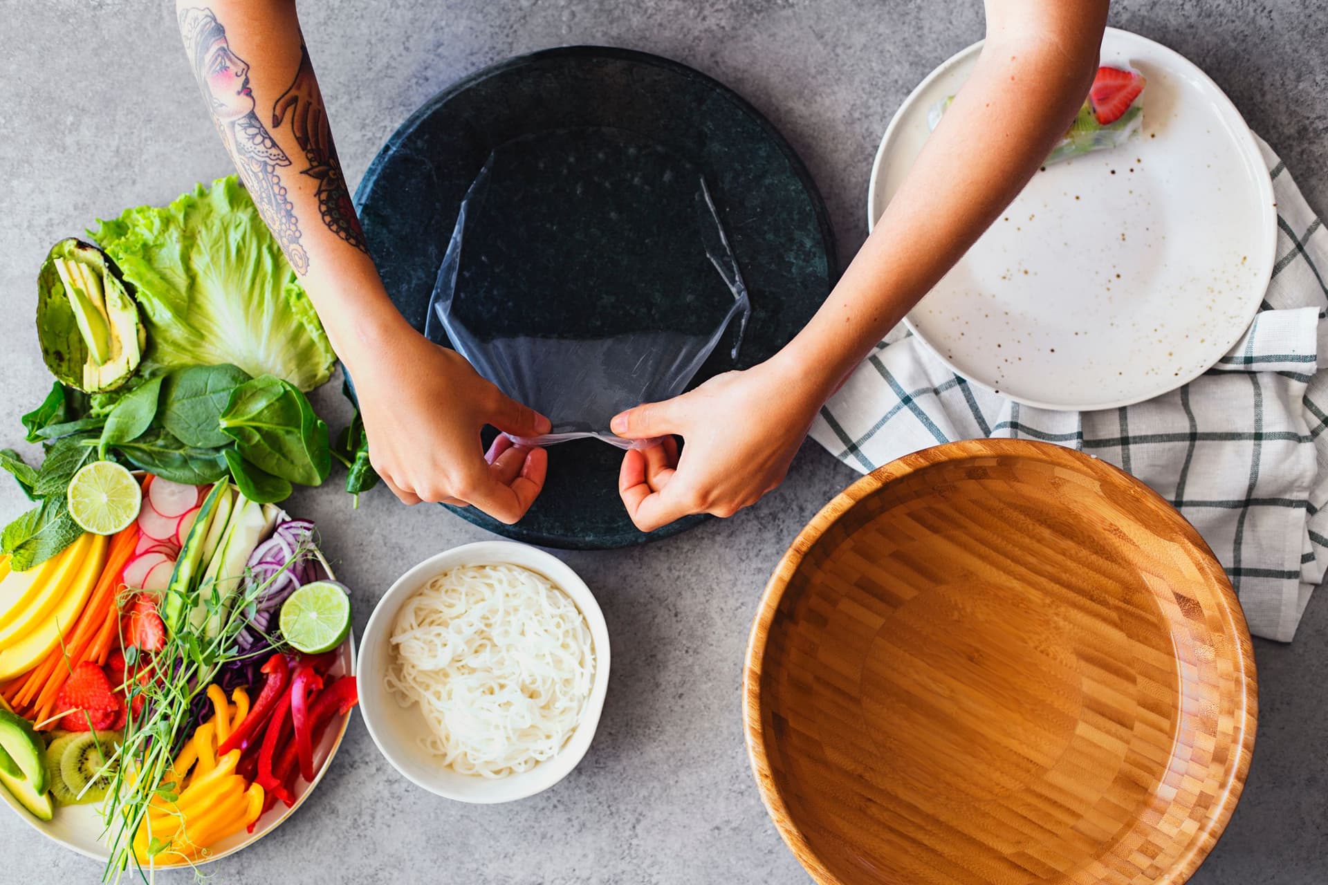 Vista de cima de mãos a preparar wraps com folhas de arroz e todos os ingredientes ao lado.