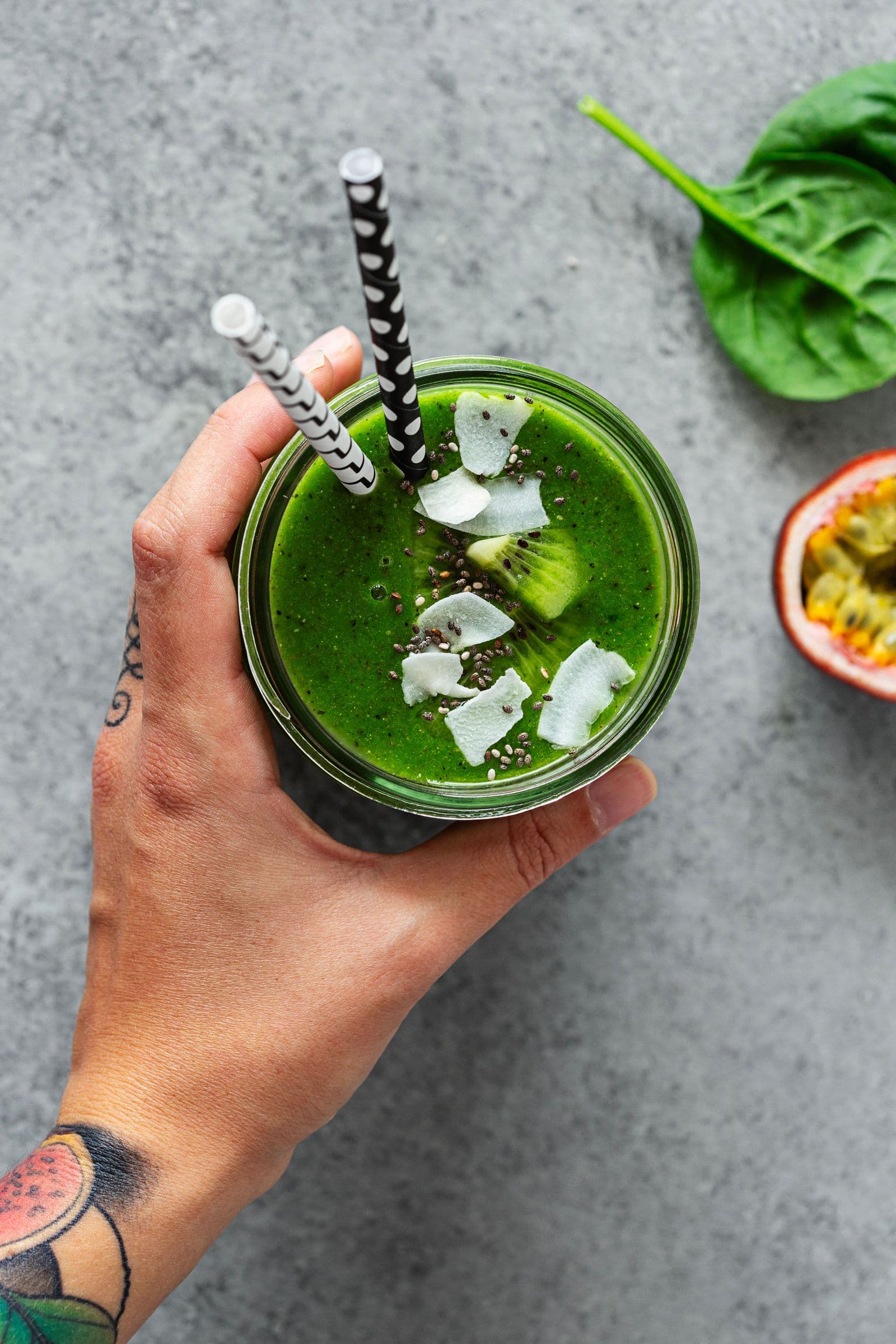 Overhead shot of a vibrant green smoothie in a jar, held by a tattooed hand, with tropical toppings
