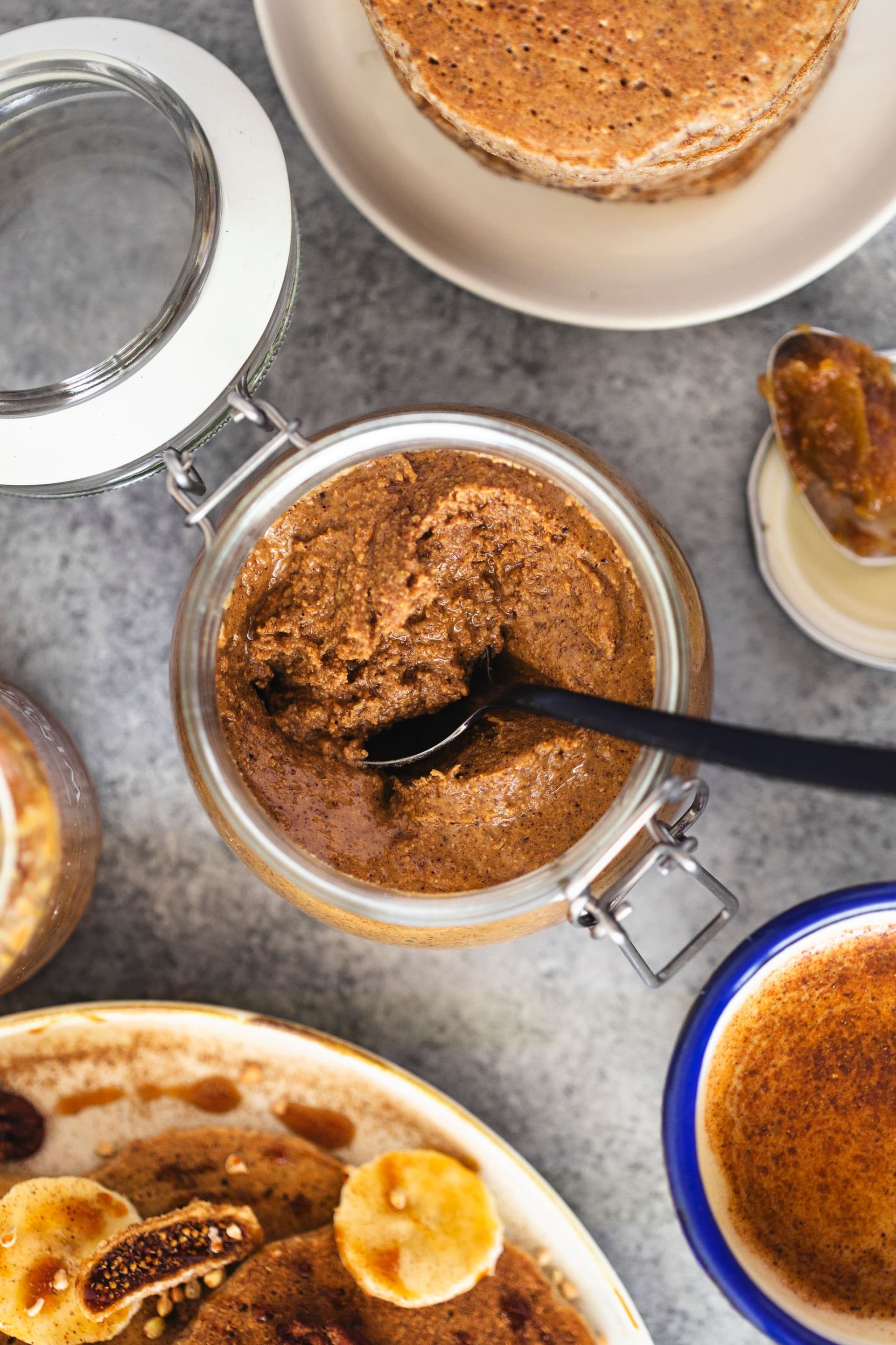 Frasco aberto de manteiga de amêndoa com especiarias chai com colher, rodeado de ingredientes para panquecas.