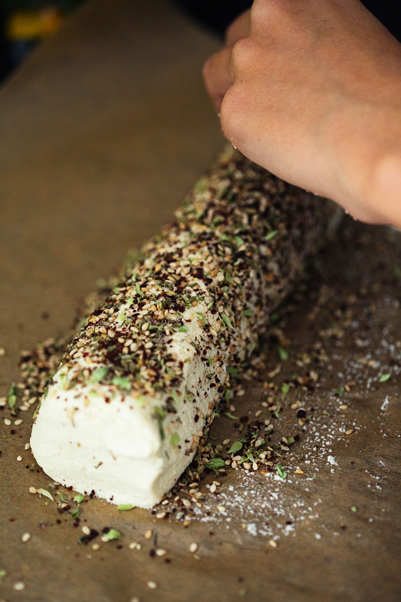 Cashew cheese log being rolled in a mix of dried herbs and spices for coating.