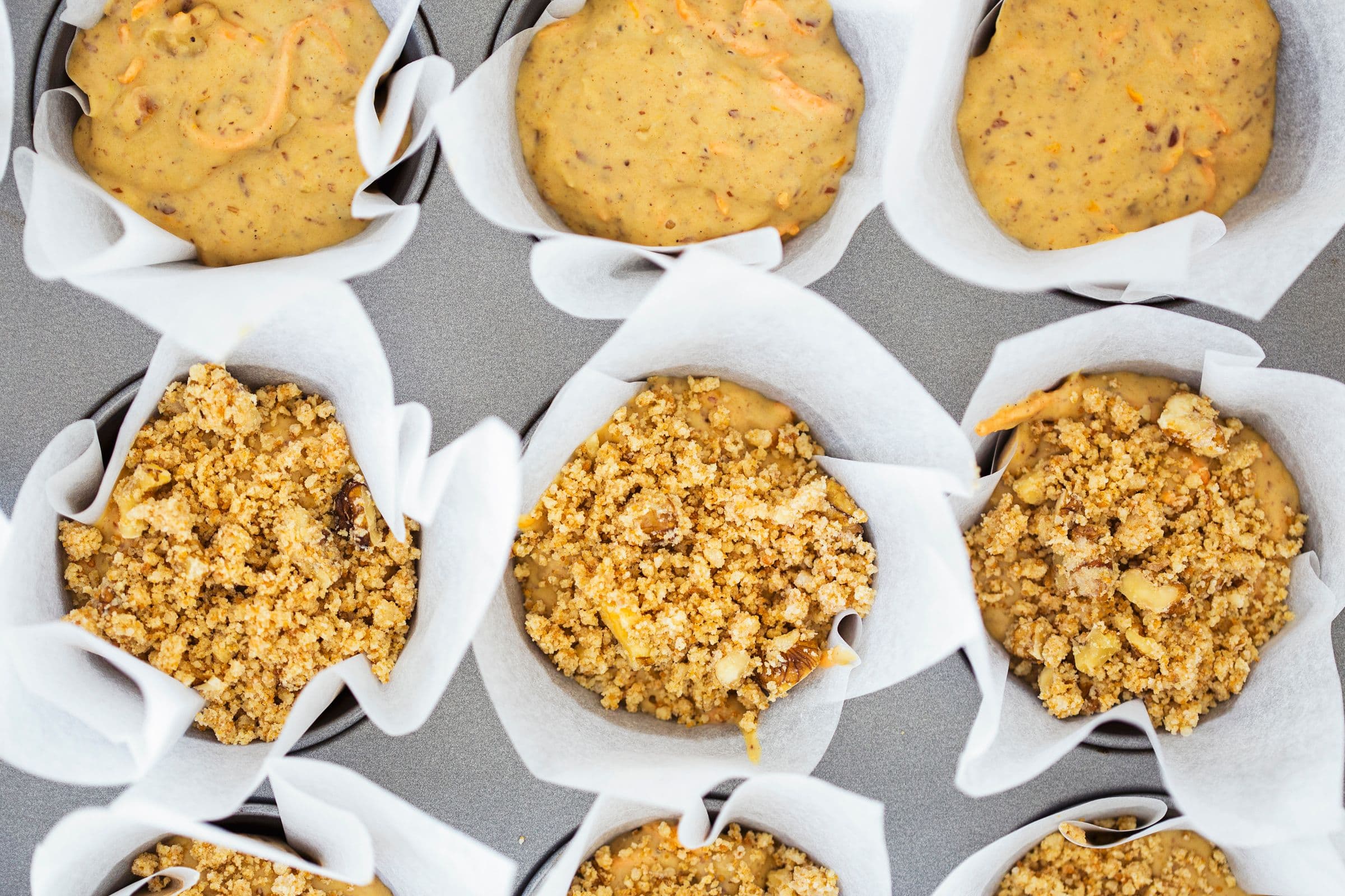 Muffin tray with batter-filled paper cases, some topped with walnut crumble.
