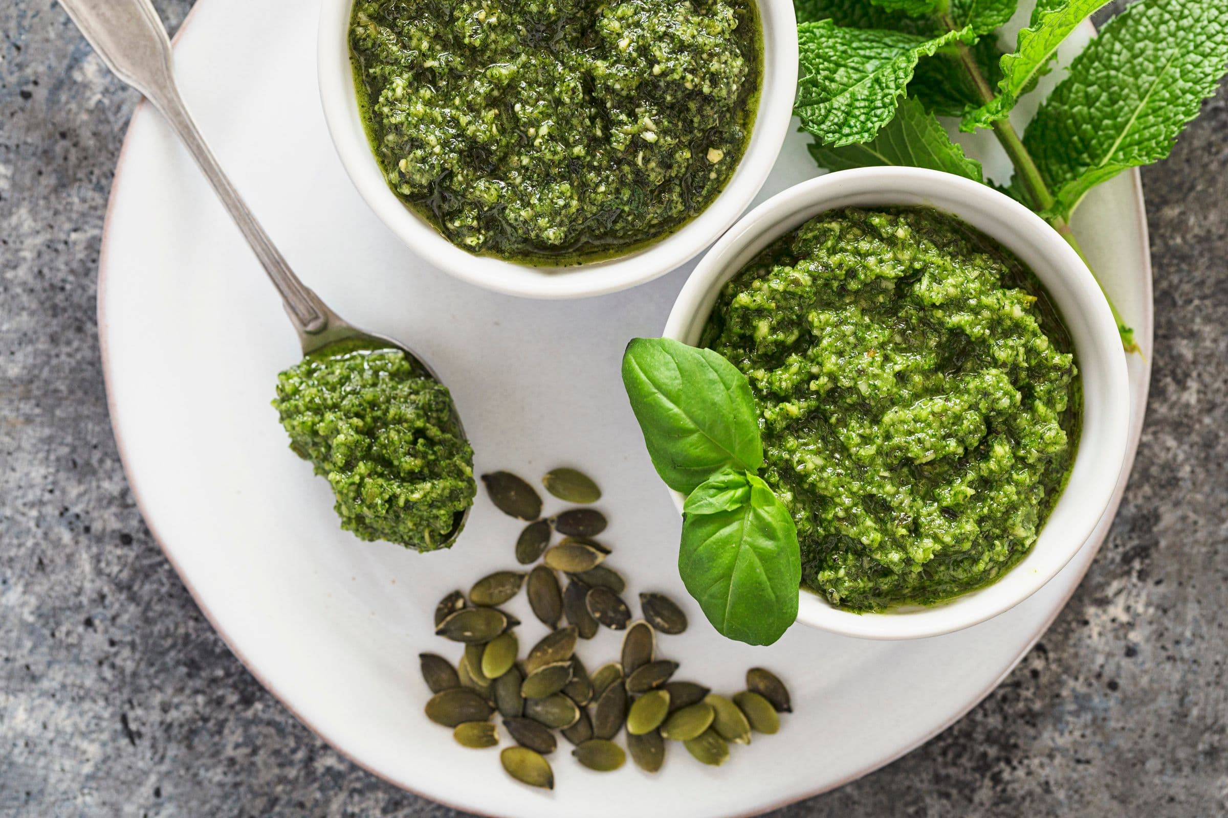 Two small bowls filled with homemade vegan pumpkin seed pesto, surrounded by fresh greens and seeds.
