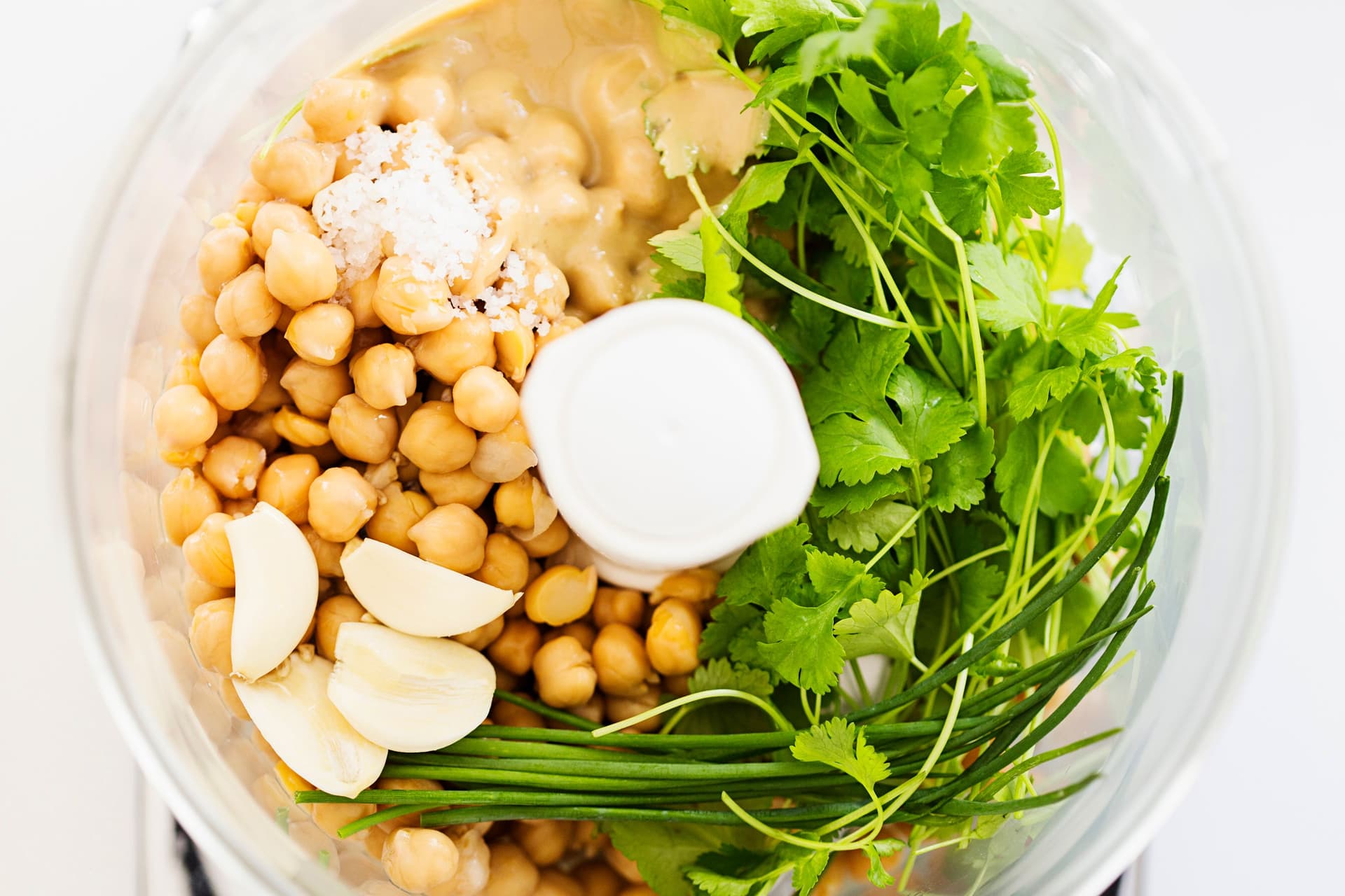 Chickpeas, garlic, tahini, and fresh herbs like coriander and chives in a food processor bowl.