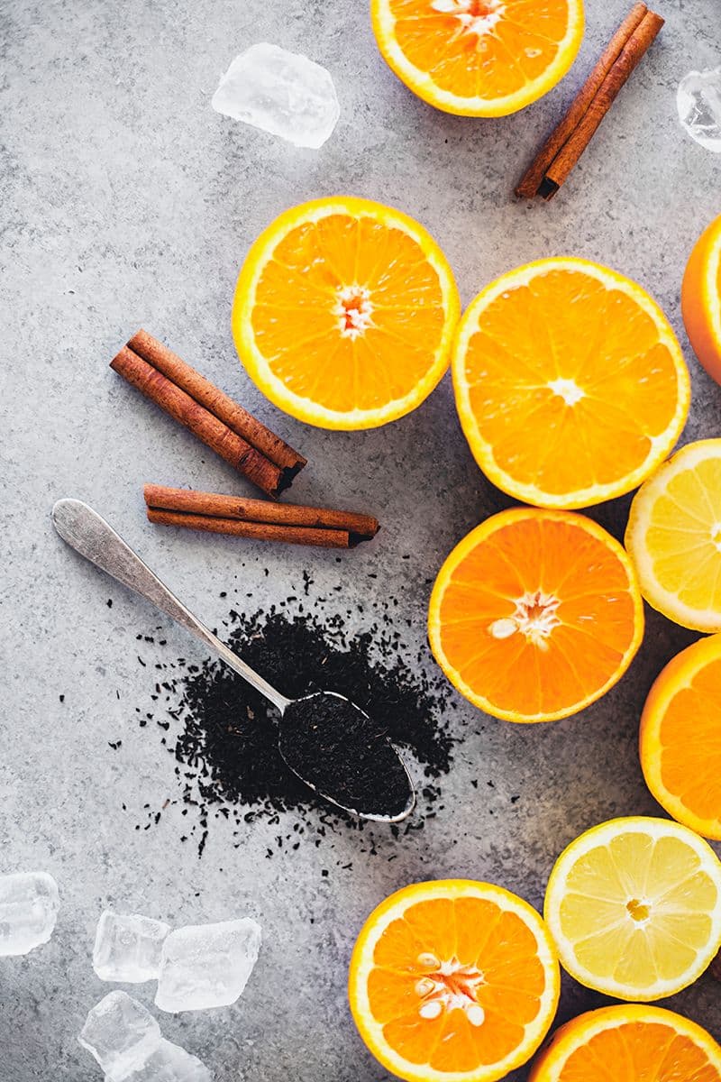 Orange and lemon slices, cinnamon sticks, loose black tea and ice cubes scattered on a surface.