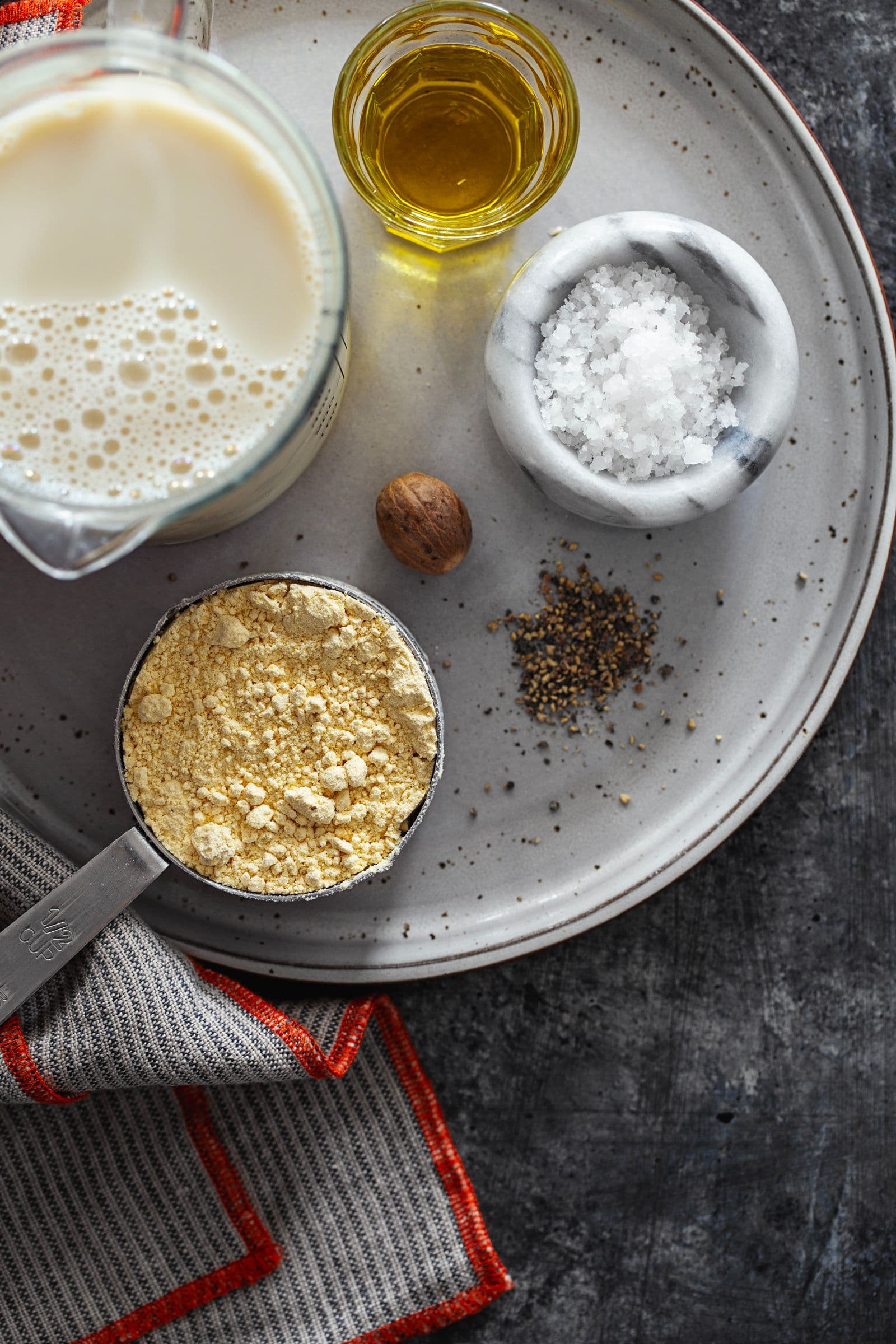 Ingredientes para béchamel vegan e sem glúten dispostos num prato: bebida vegetal, farinha de grão, azeite, sal, noz-moscada e pimenta
