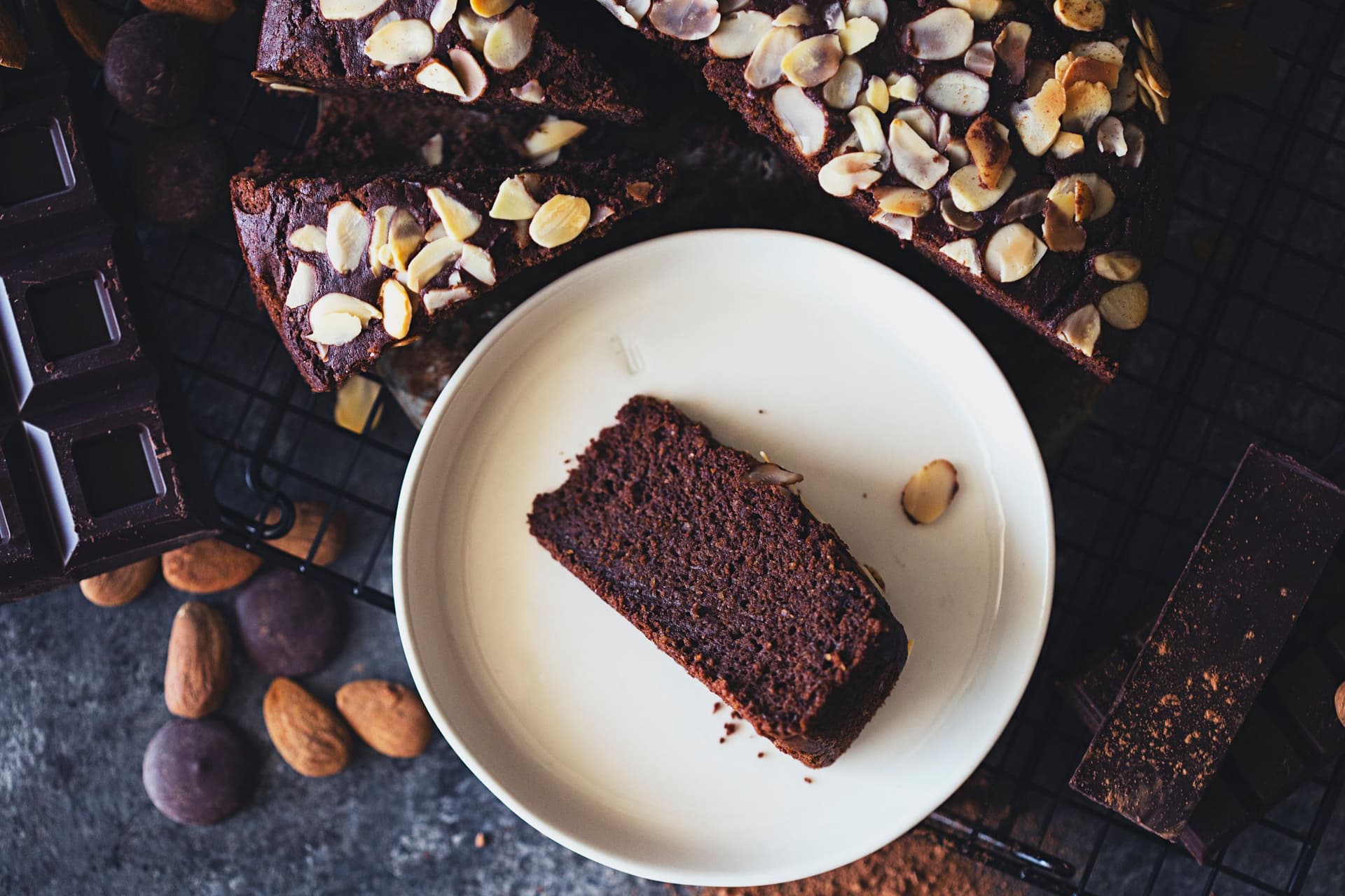 Uma fatia de bolo de chocolate com amêndoa servida num prato branco com chocolate e amêndoas ao lado.
