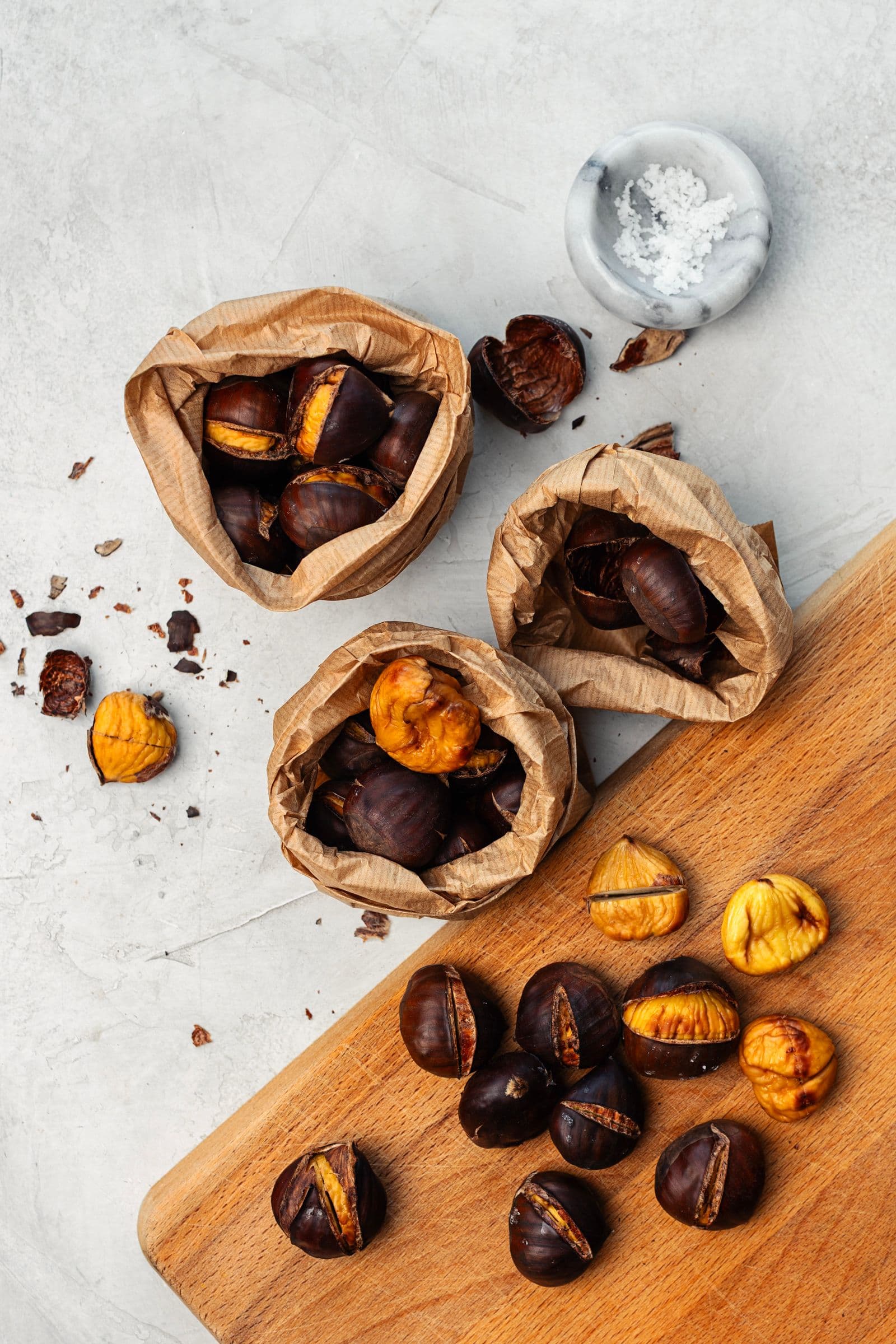 Three brown paper bags filled with roasted chestnuts, with loose shells and opened chestnuts scattered on the cutting board.
