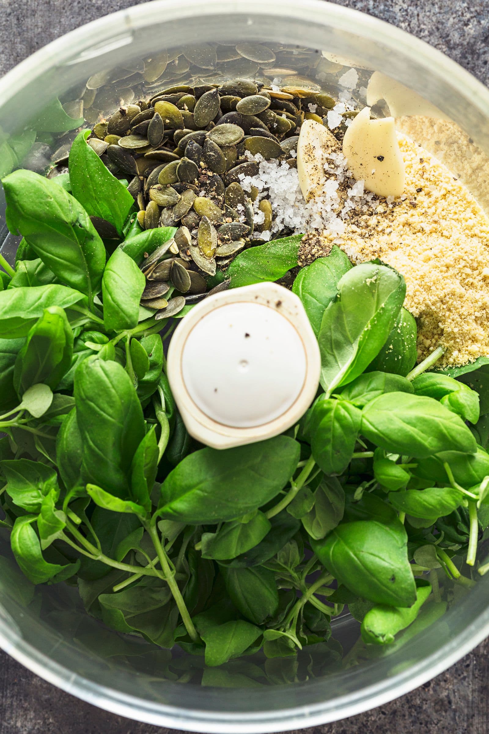 Pumpkin seeds, garlic, olive oil and green herbs placed in a food processor bowl before blending.