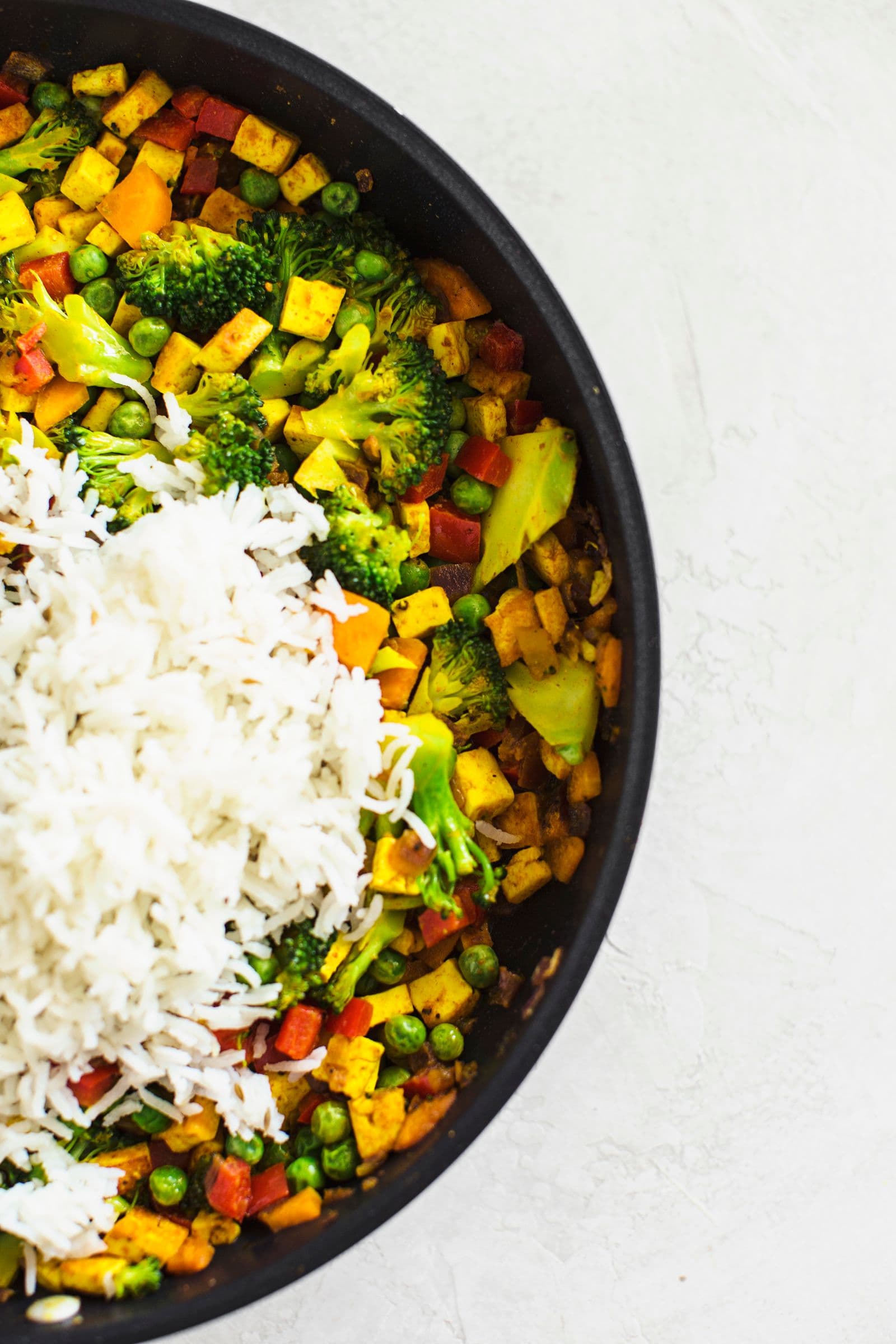 Arroz cozido a ser adicionado a legumes salteados numa frigideira preta.