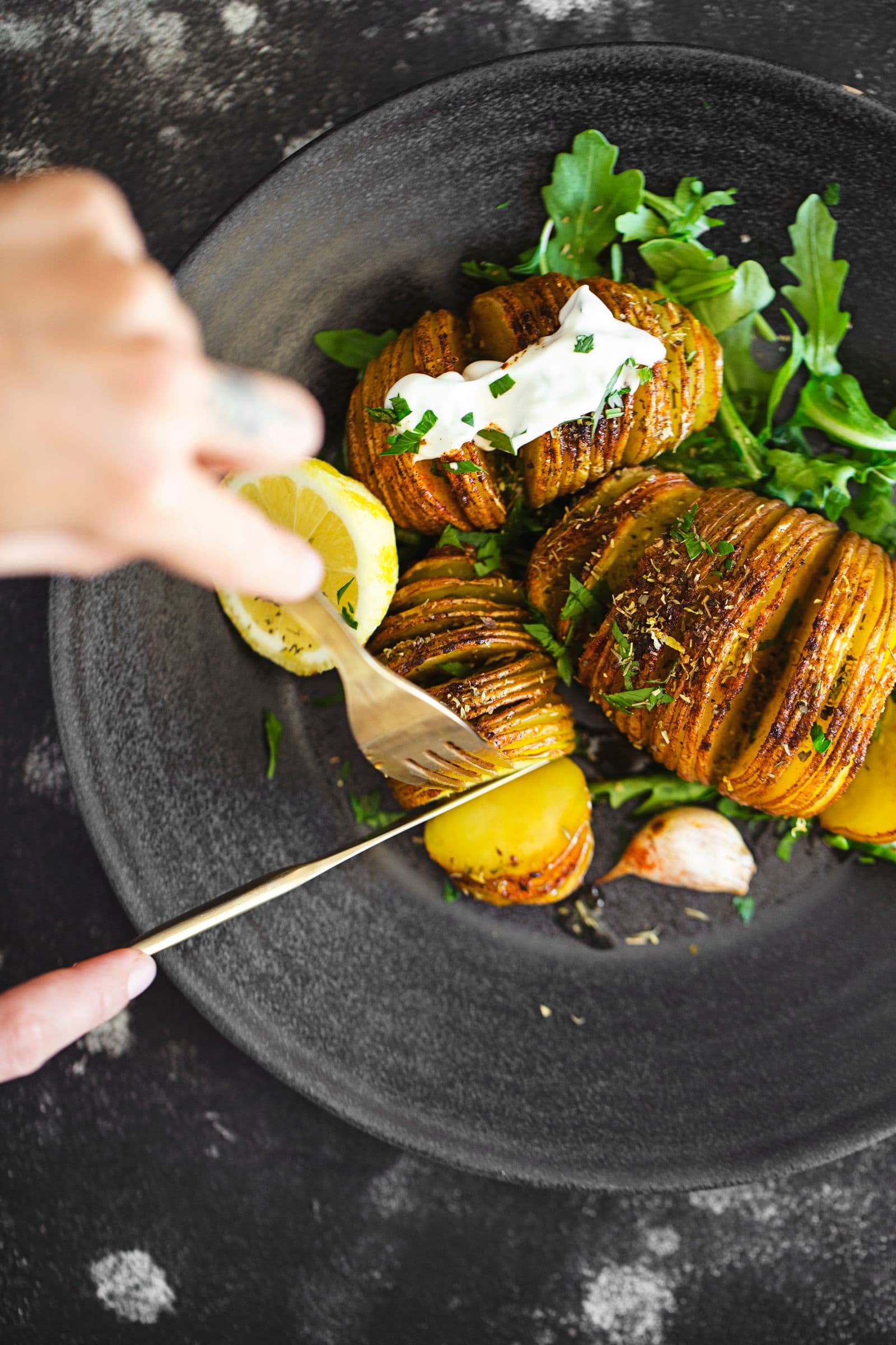 Dark plate with crispy Hasselback potatoes, arugula, lemon wedge and mayo as a hand cuts a piece.