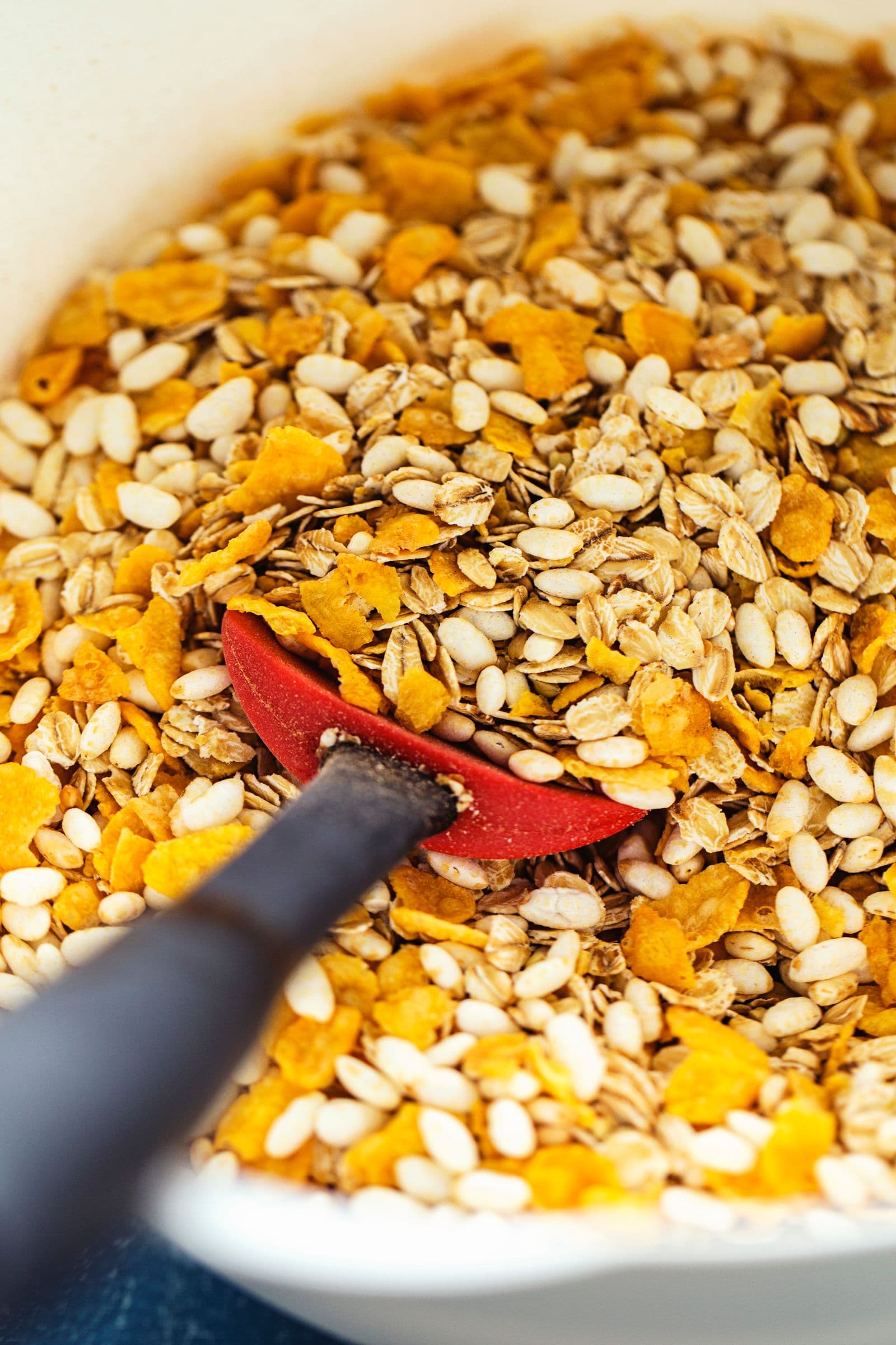 A spatula mixing oats, cornflakes and seeds in a large bowl.