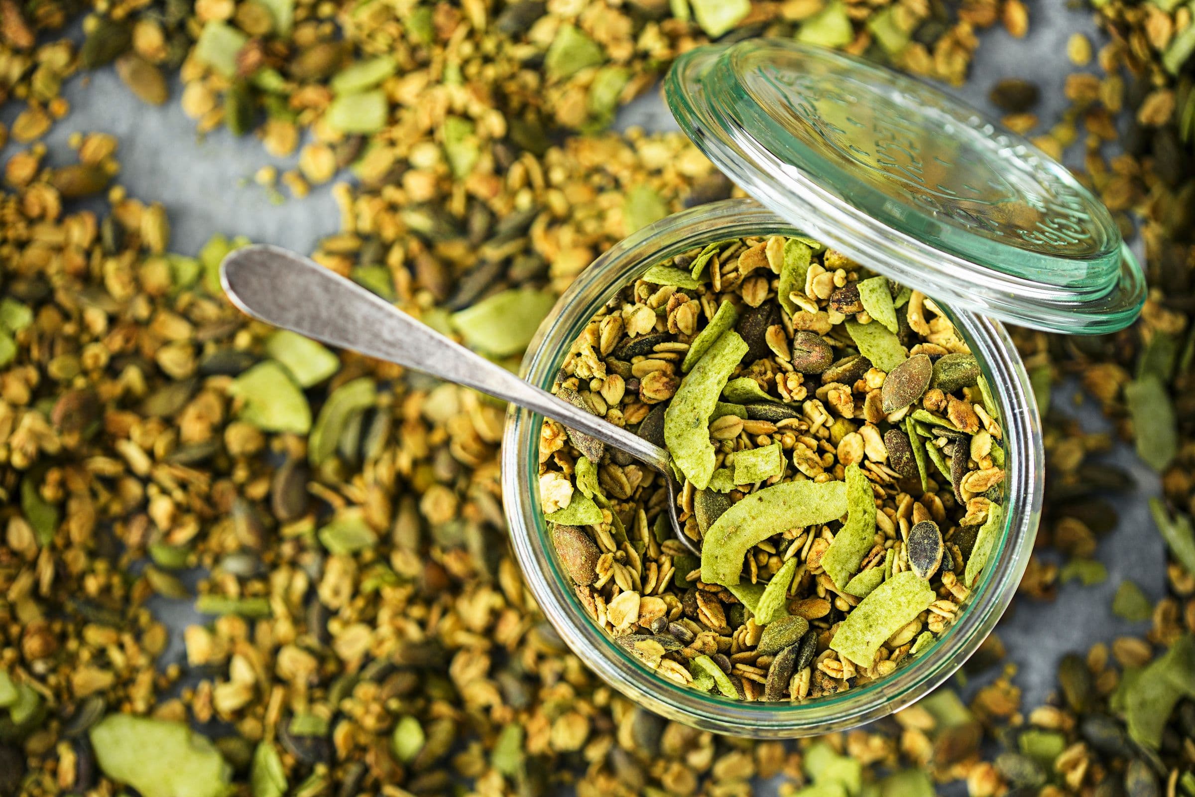 Frasco de vidro com granola de coco e lima e colher apoiada no interior.