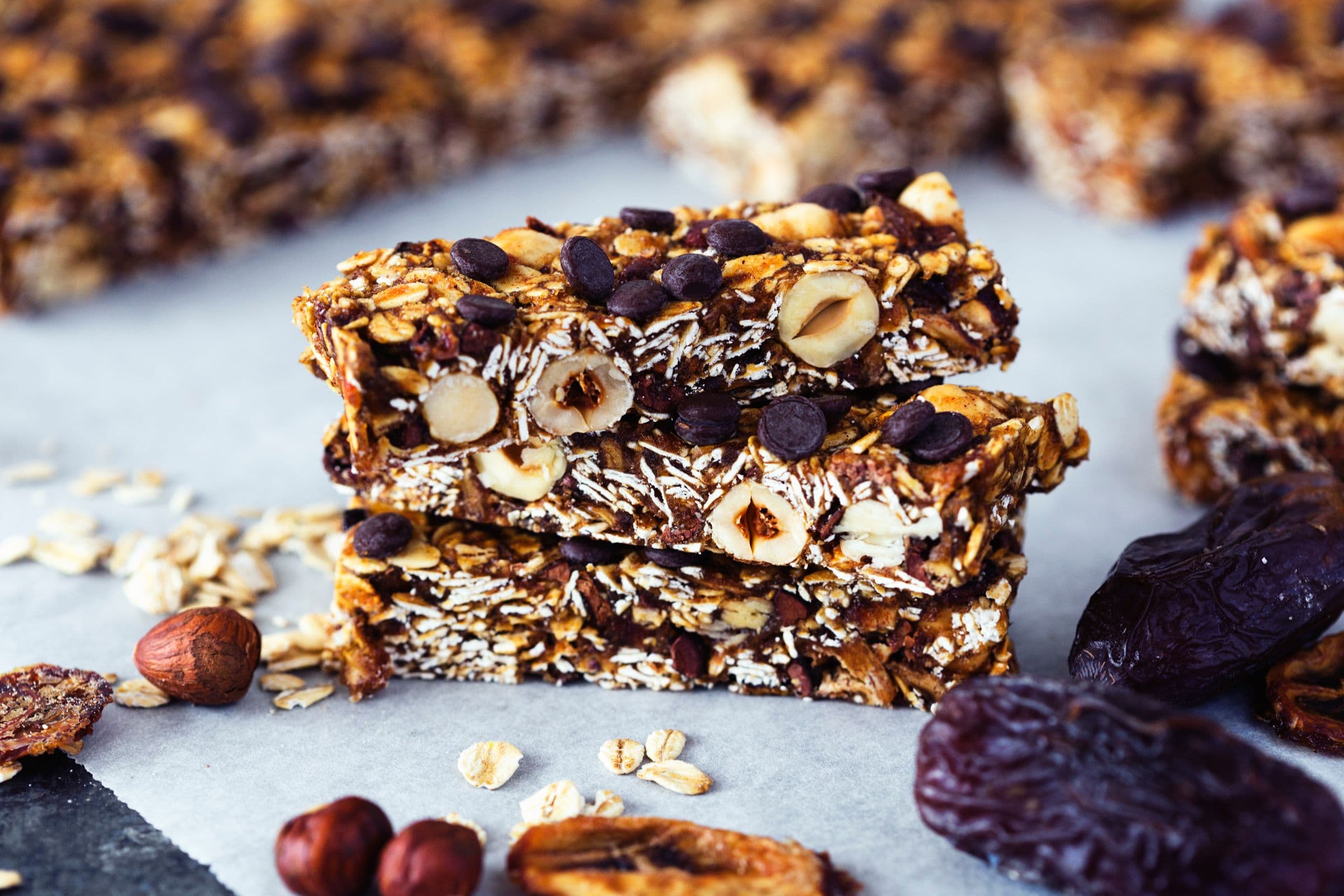 Barras de granola empilhadas com avelãs, chips de banana e pepitas de chocolate sobre papel vegetal.