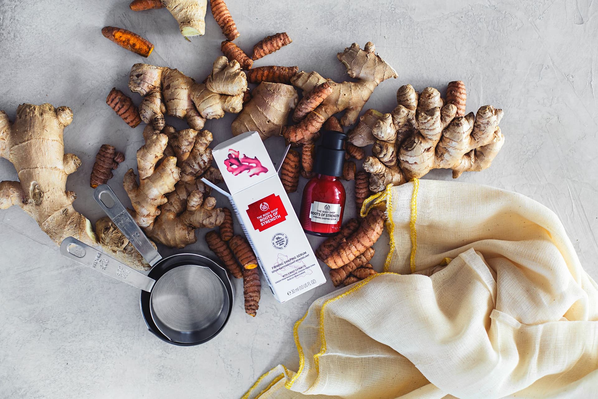 Raízes frescas de gengibre e curcuma com um frasco de sérum da The Body Shop sobre fundo claro.