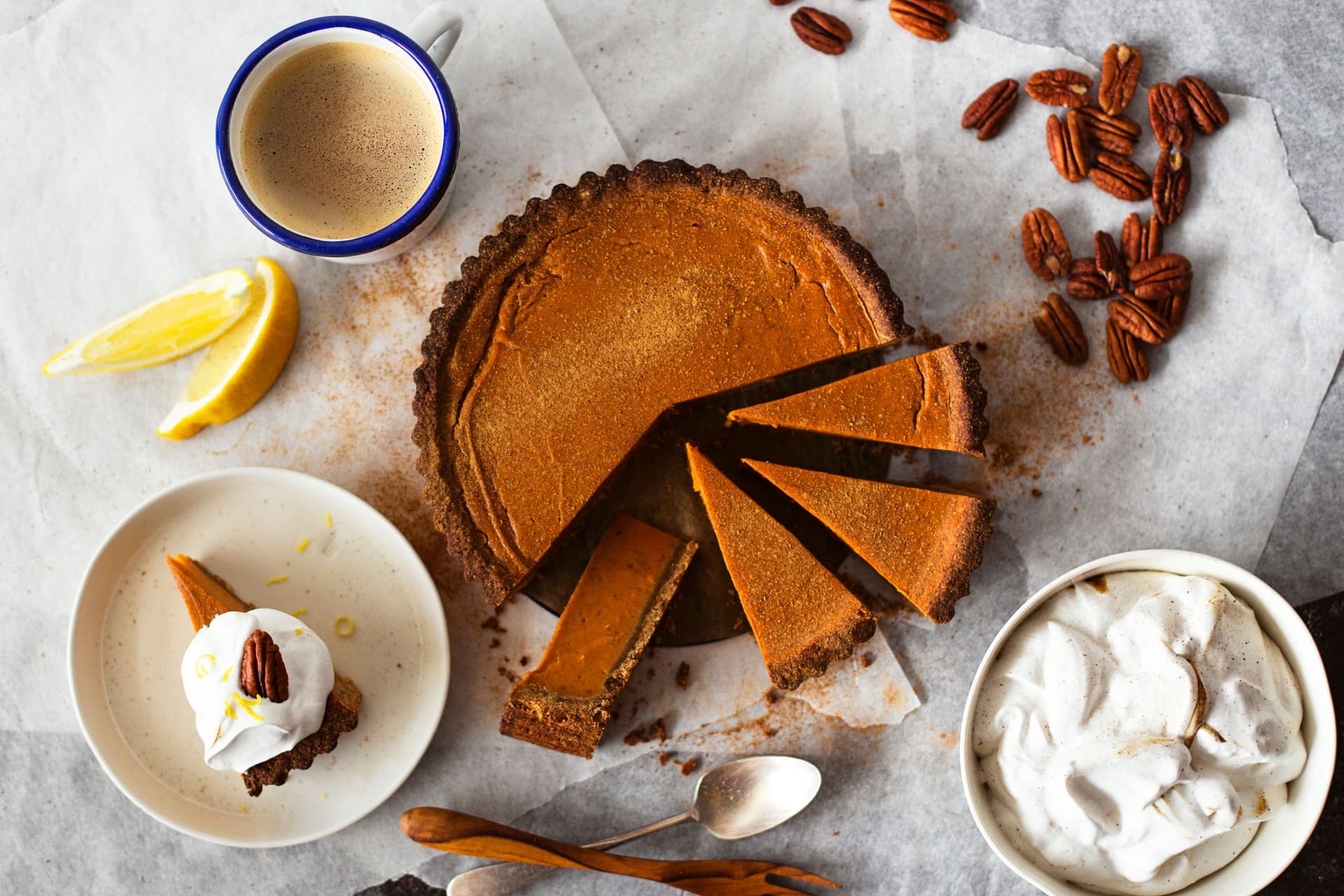 Tarte de batata-doce com uma fatia servida num prato, coberta com chantilly vegan e nozes pecan, rodeada de chávenas de café.