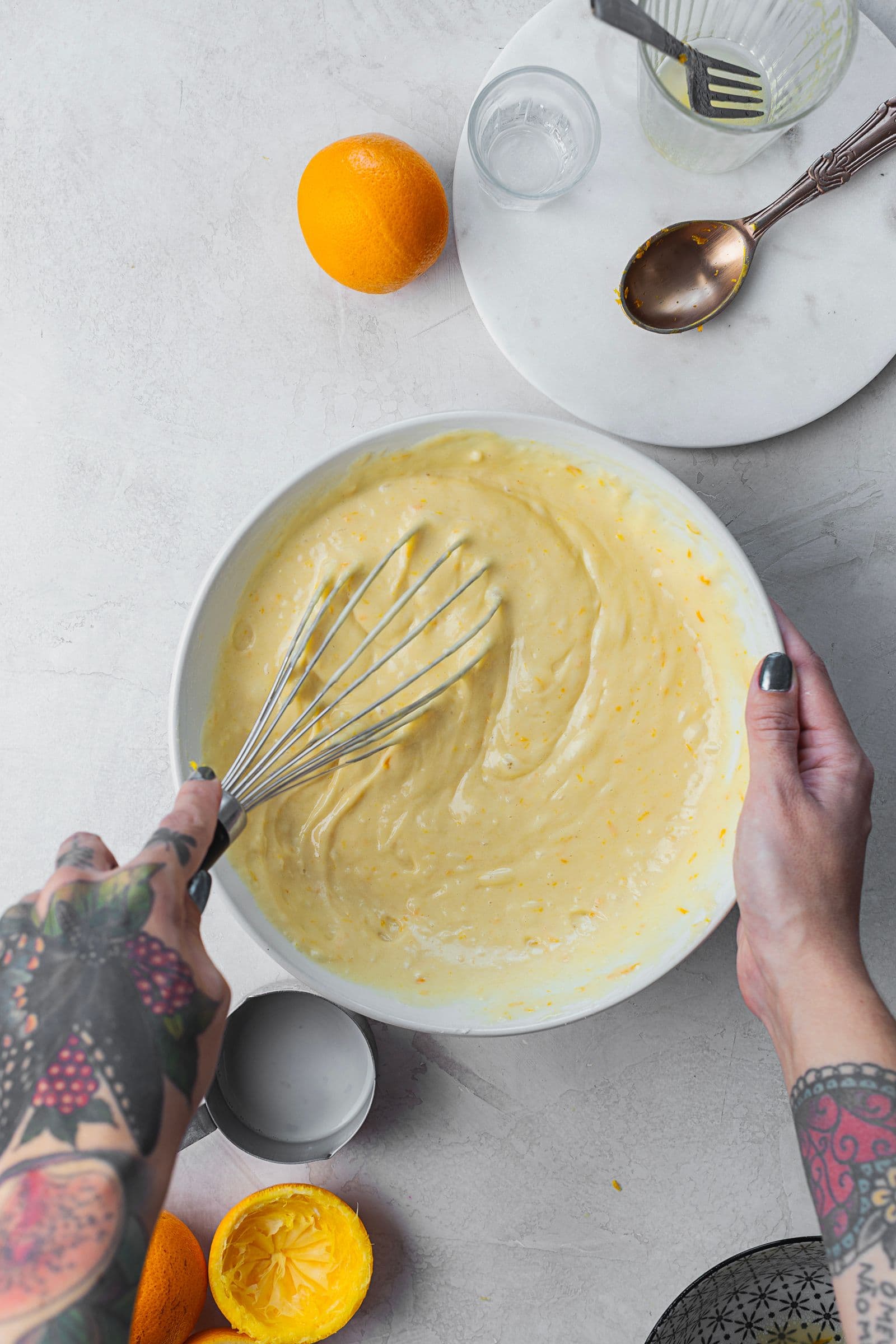 Mãos a misturar a massa do bolo de laranja com um fouet numa taça branca, rodeada de laranjas e utensílios de cozinha.