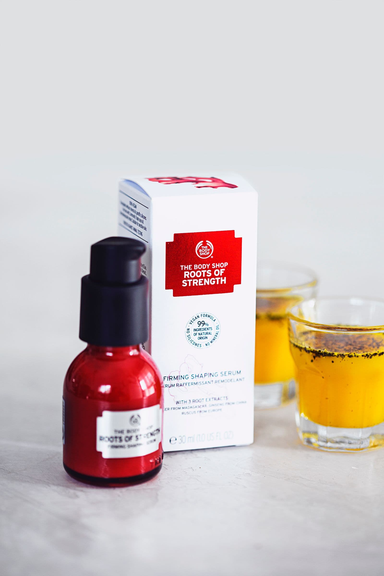 Pequeno frasco de sérum da The Body Shop ao lado de dois copos com xarope de gengibre e curcuma sobre fundo branco.