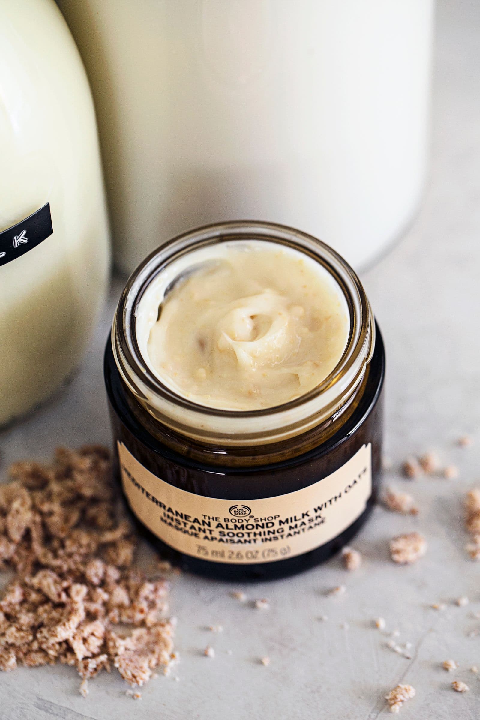 Frasco aberto de máscara hidratante de amêndoa e aveia da The Body Shop, com textura cremosa visível, ao lado de leite vegetal em garrafas de vidro.