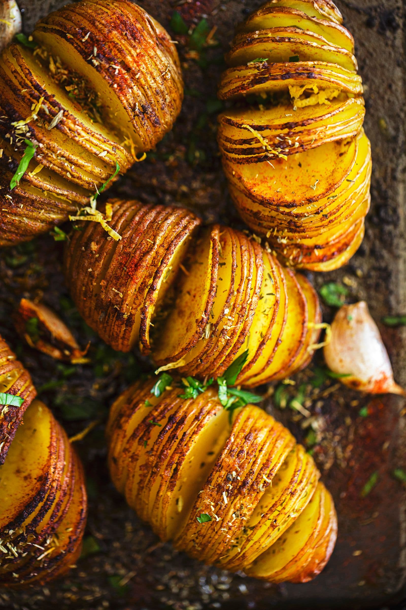 Close view of a seasoned Hasselback potato on a dark baking tray with roasted garlic.