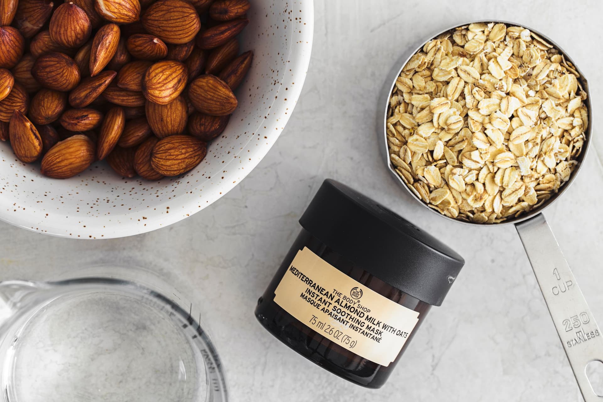 Ingredientes para leite vegetal: tigela com amêndoas, medidor com flocos de aveia e frasco de máscara hidratante de amêndoa e aveia da The Body Shop.