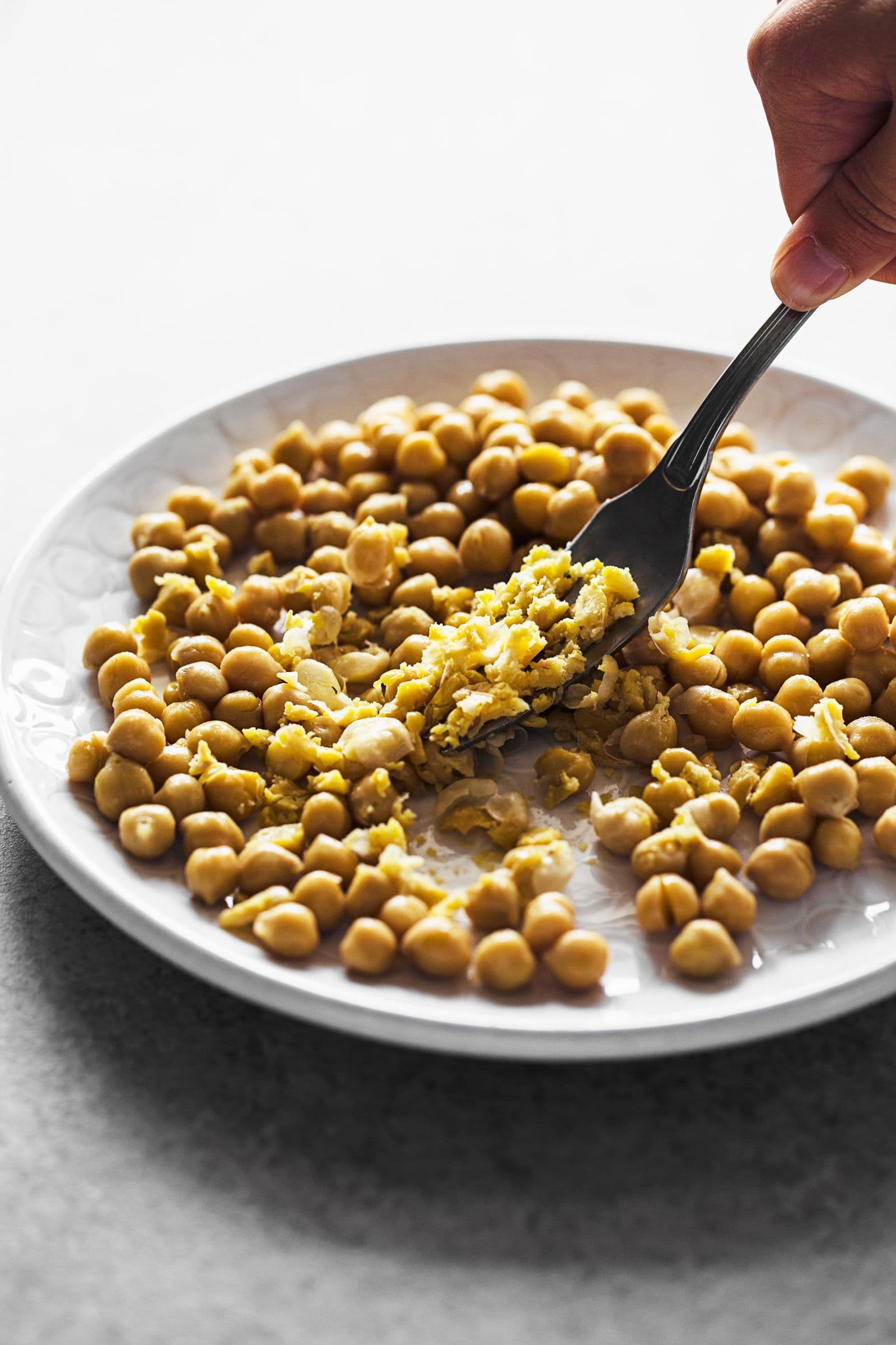 Hand using a fork to smash chickpeas into a paste on a white dish.
