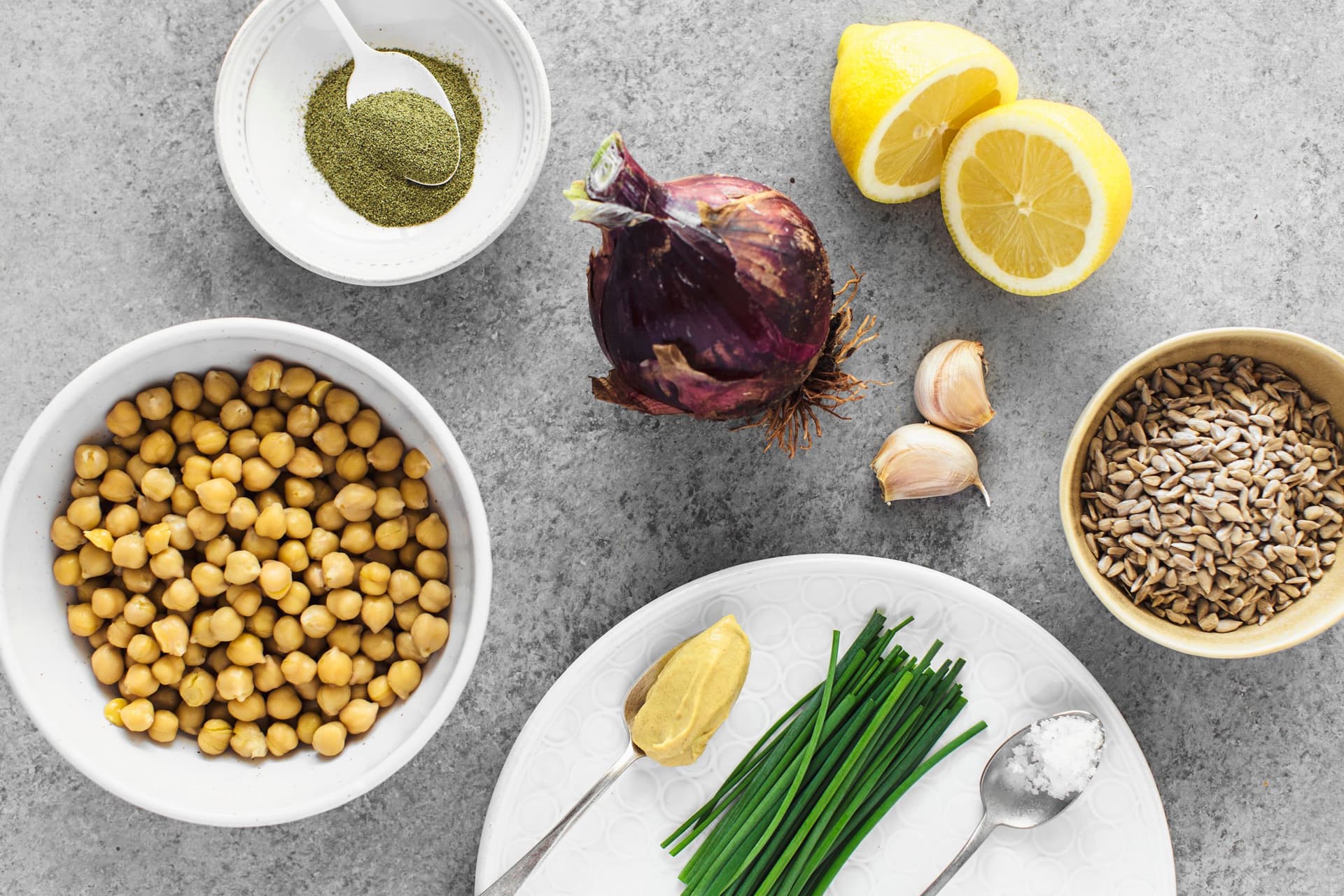 Grão-de-bico, sementes de girassol, limão, cebola, alho, cebolinho e mostarda dispostos sobre fundo branco.