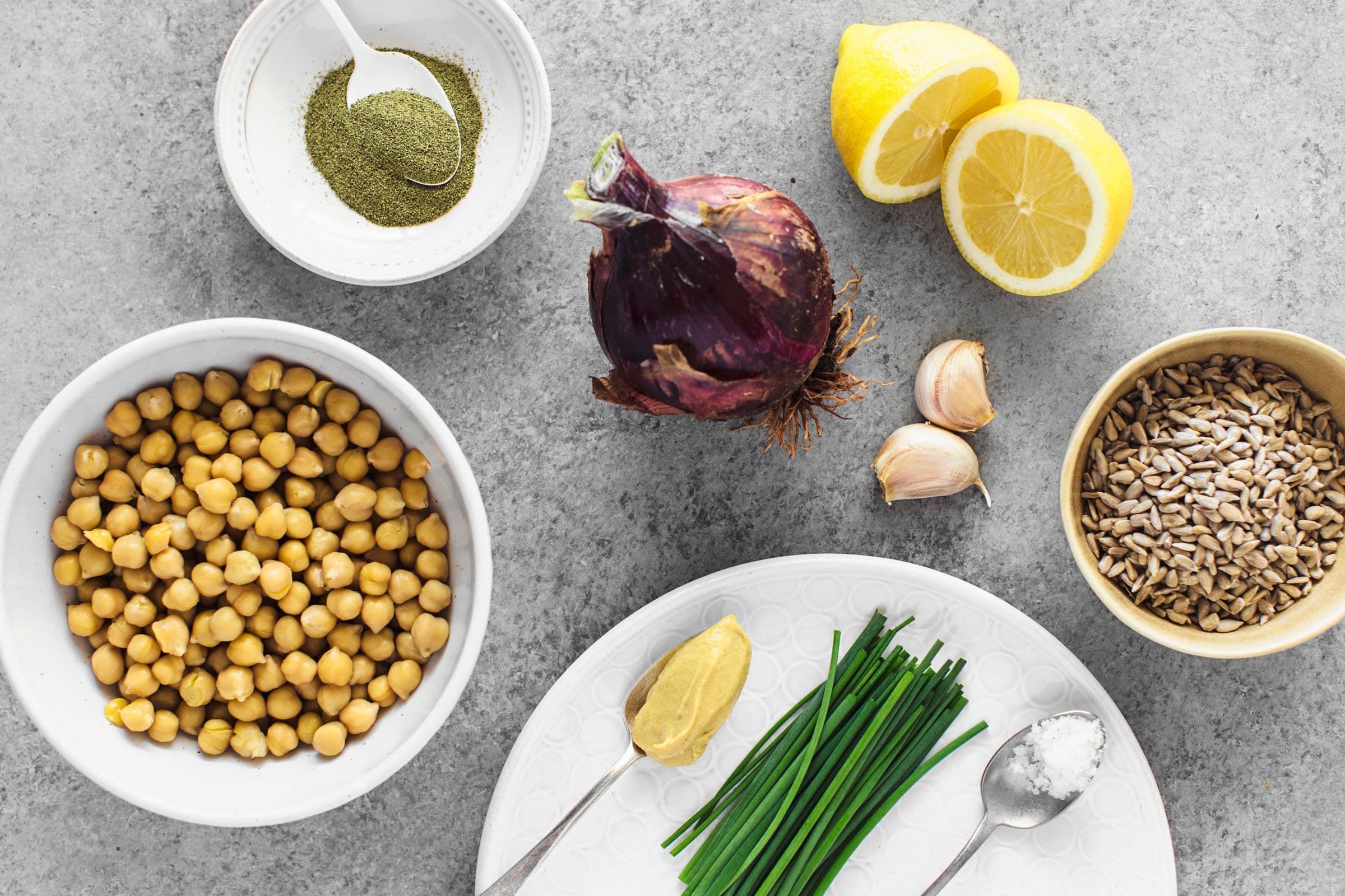 Chickpeas, sunflower seeds, lemon, onion, garlic, chives and mustard arranged on a light grey background.