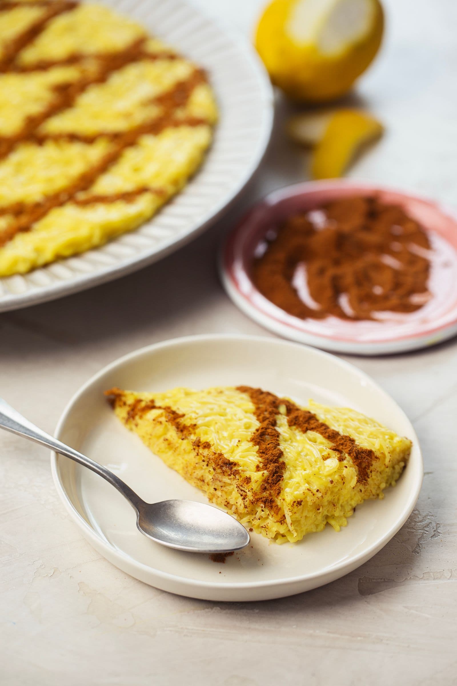 Slice of vegan Aletria on a small white plate with a spoon, with the rest of the dessert and cinnamon in the background.