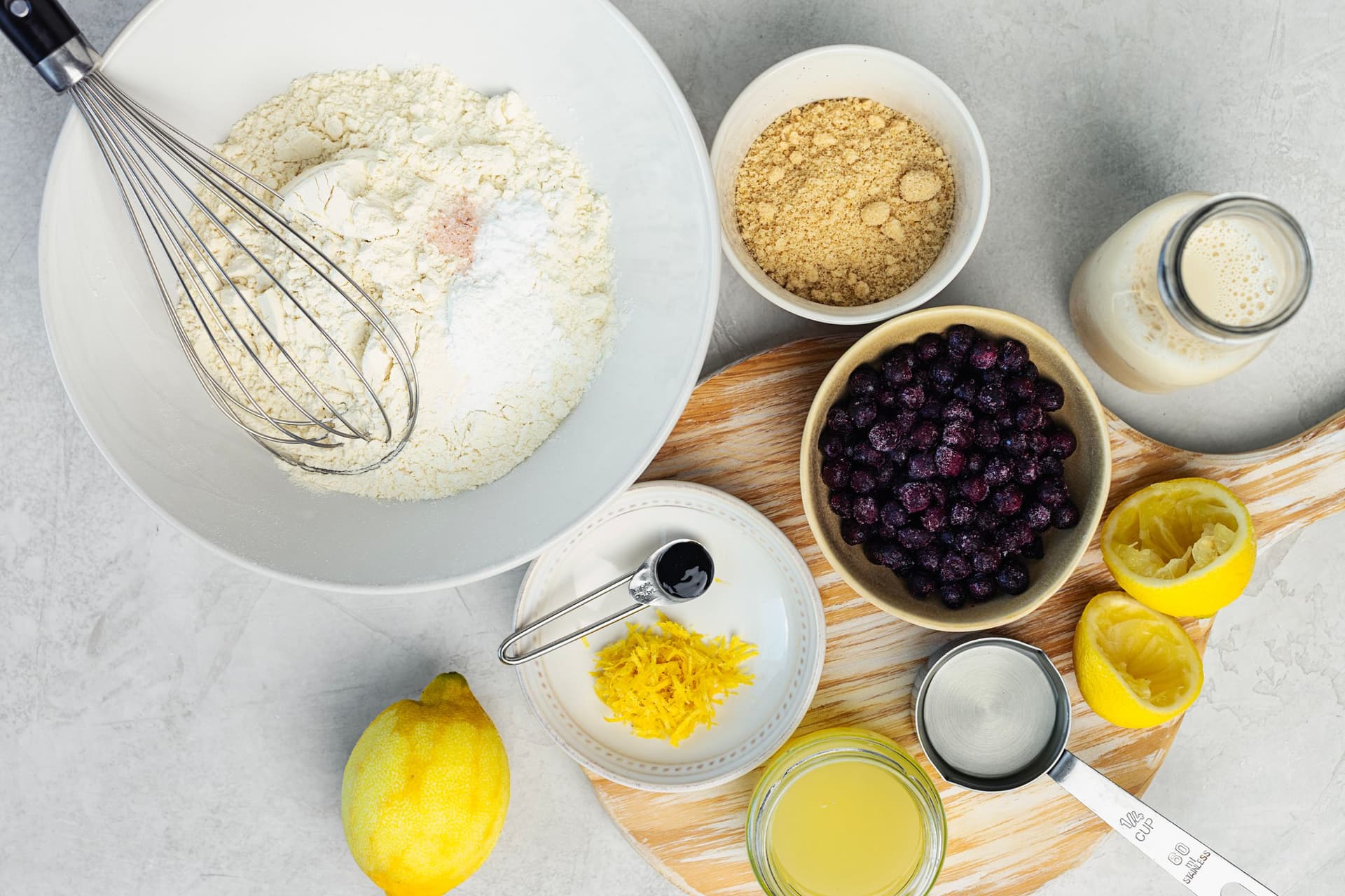 Ingredientes para queques de limão e mirtilos com crumble, dispostos numa bancada clara, incluindo farinha, raspa de limão, mirtilos e bebida vegetal.