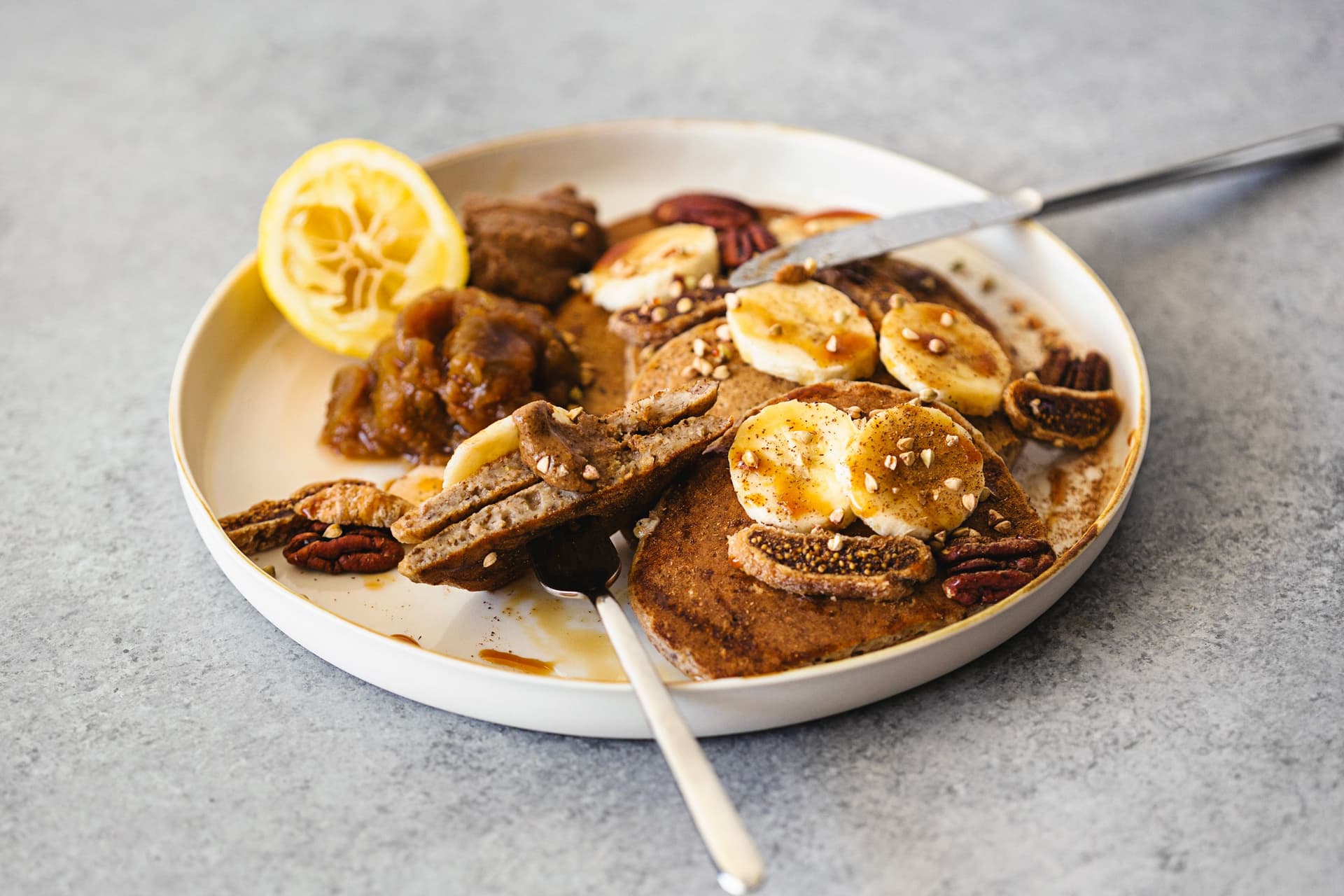 Individual portion of pancakes with lemon half, fig jam and banana on a round white plate.