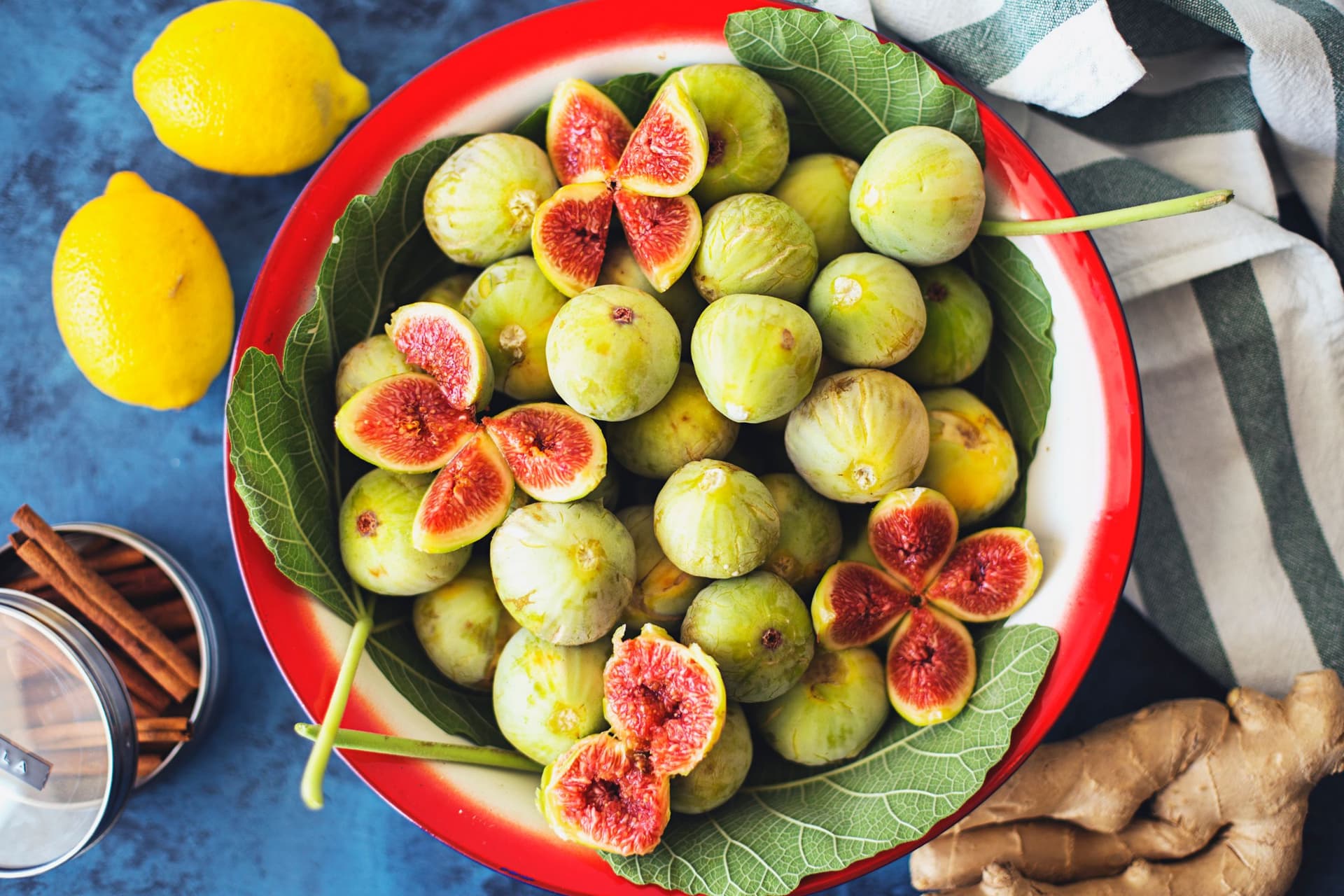 Red bowl with halved figs, whole green figs, ginger and lemon on a striped cloth.
