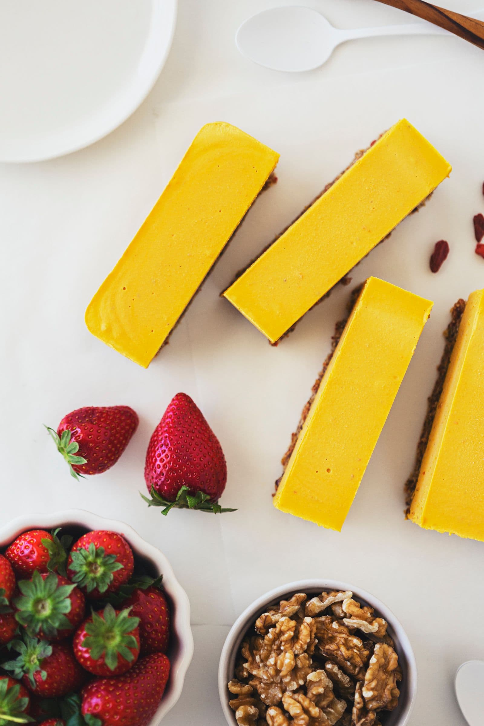 Barras de cheesecake cortadas, com taças de nozes e morangos ao lado.